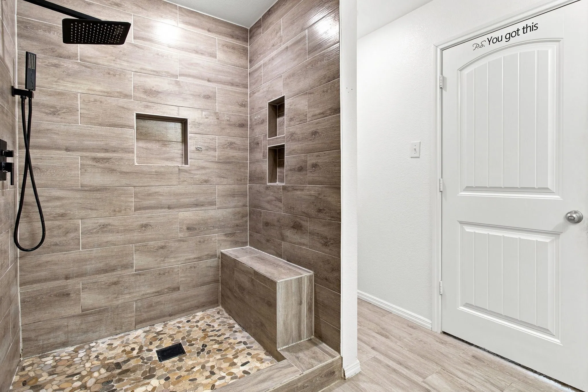 Walk-in shower featuring wood-finish tile walls, built-in shelving, a shower bench, and a river rock floor