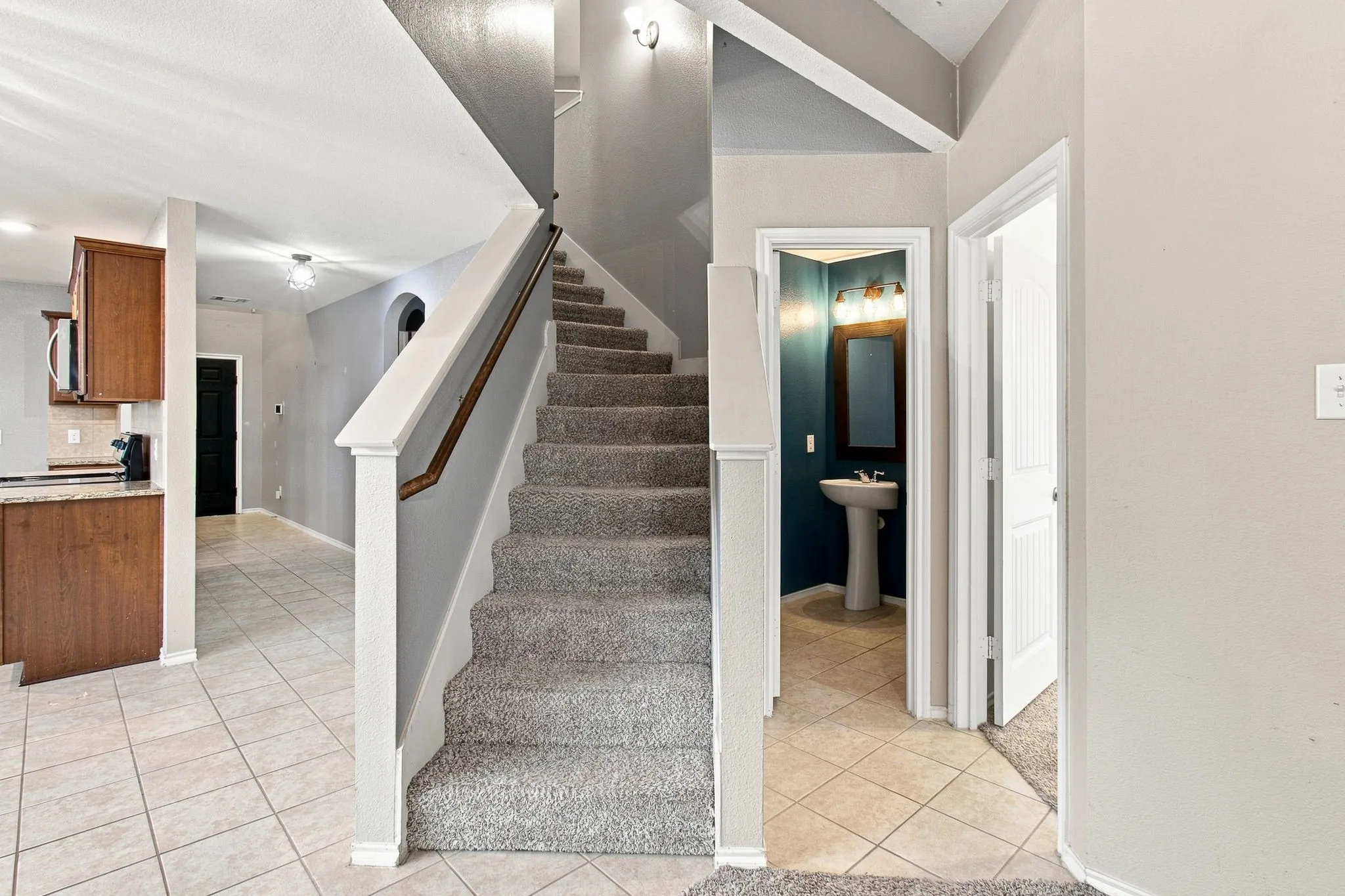 Carpeted staircase with wood handrail, pedestal sink, wall-mounted mirror, and tile flooring