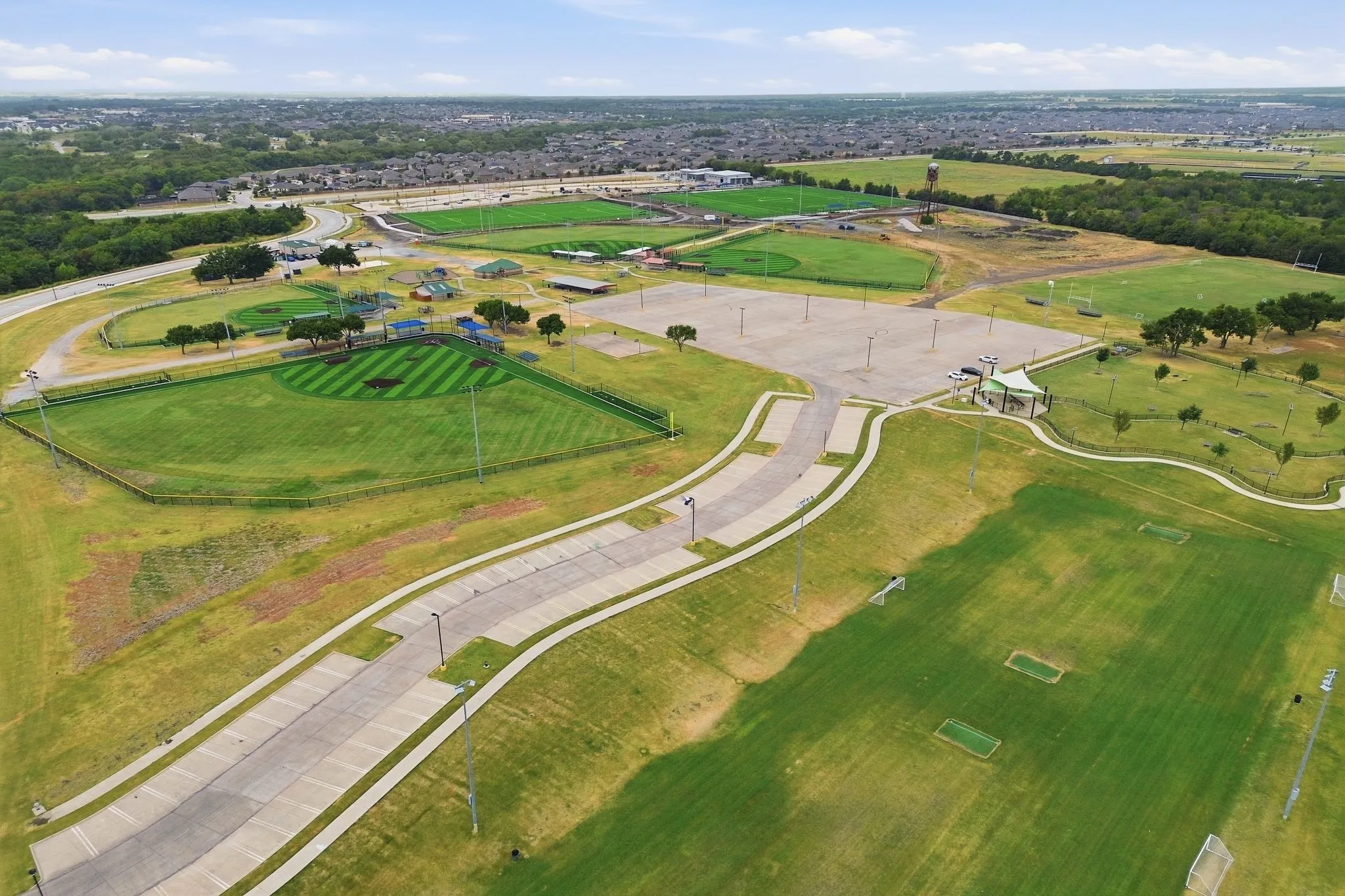 Expansive community sports complex featuring multiple athletic fields, a baseball diamond, paved pathways, a large parking area, and a central covered pavilion