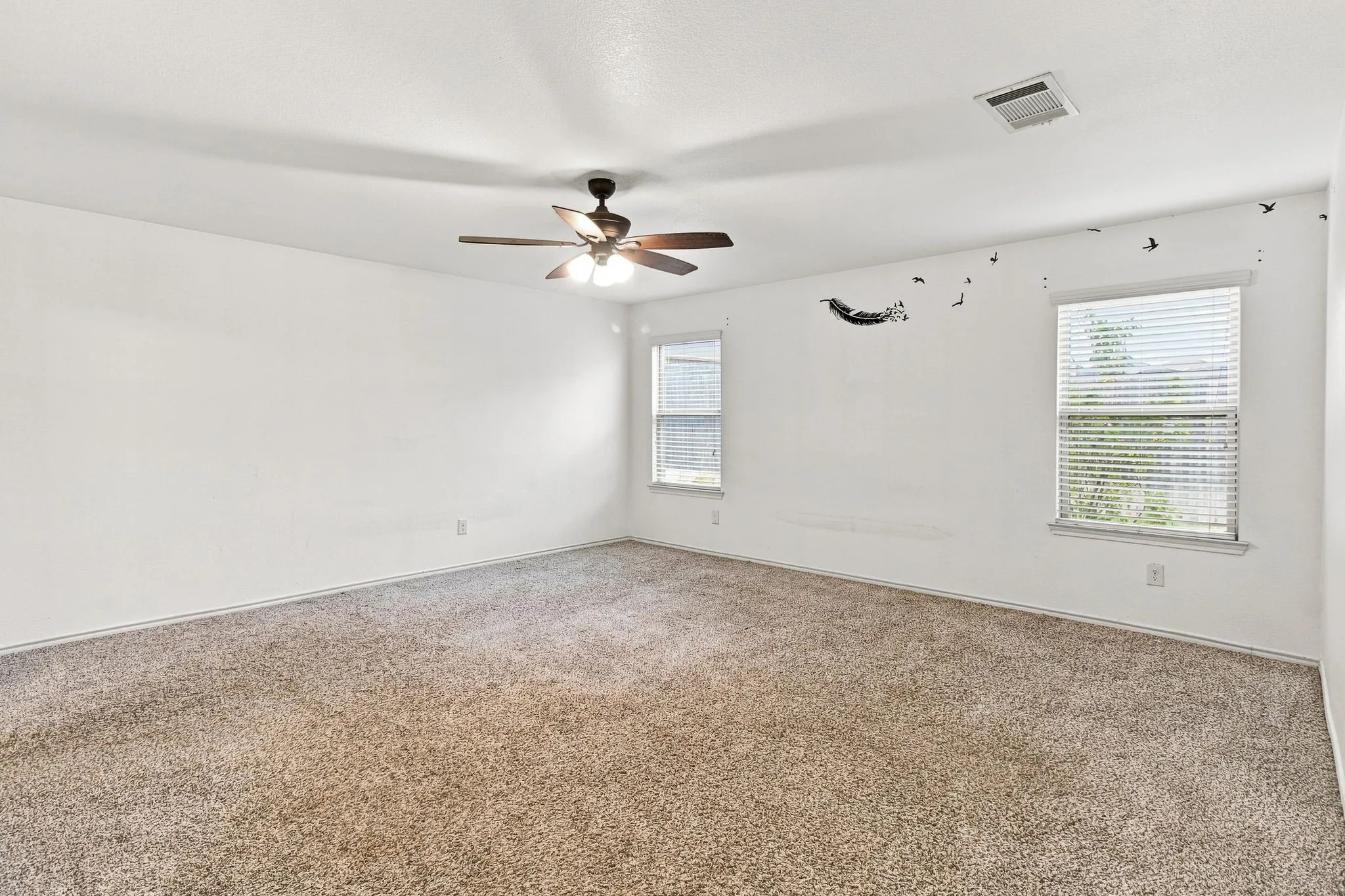 Spacious room featuring carpet flooring, two windows with blinds, a ceiling fan with integrated lighting, and white walls