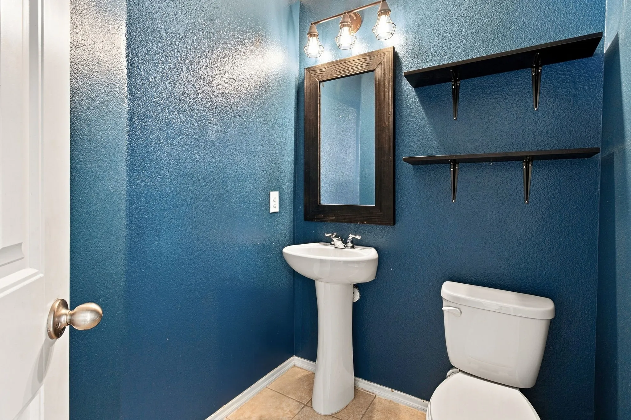 Pedestal sink with chrome faucet, wall-mounted mirror with dark frame, overhead vanity lighting, two wall-mounted shelves, and tile flooring