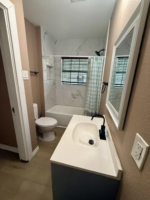 Wood-finish flooring, white vanity with integrated sink, black single-handle faucet, white framed mirror, and white tiled shower surround with a built-in niche