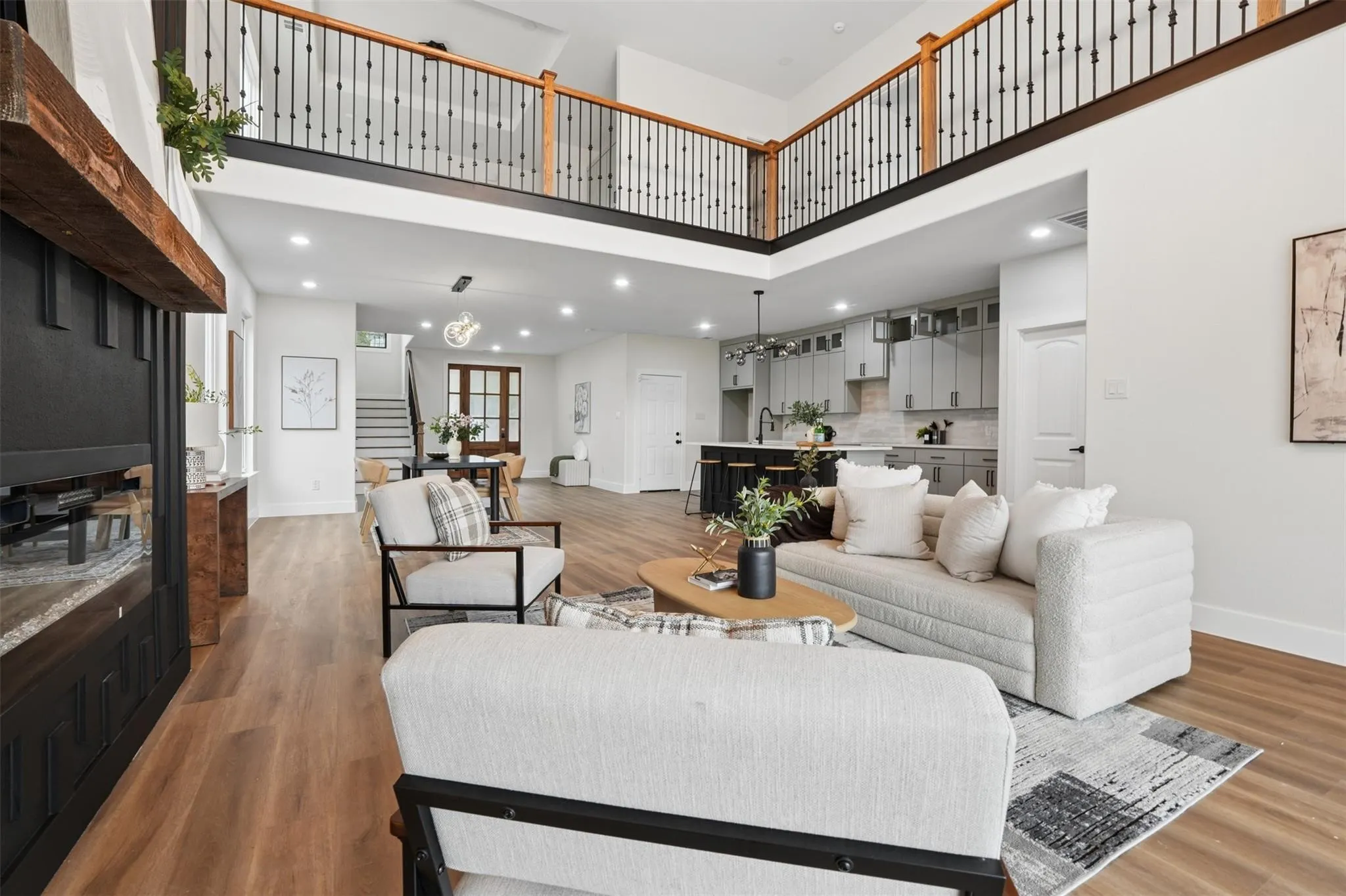 Two-story living area with wood-finish flooring, a contemporary fireplace, and an open concept kitchen featuring light gray cabinetry and a dark island with seating
