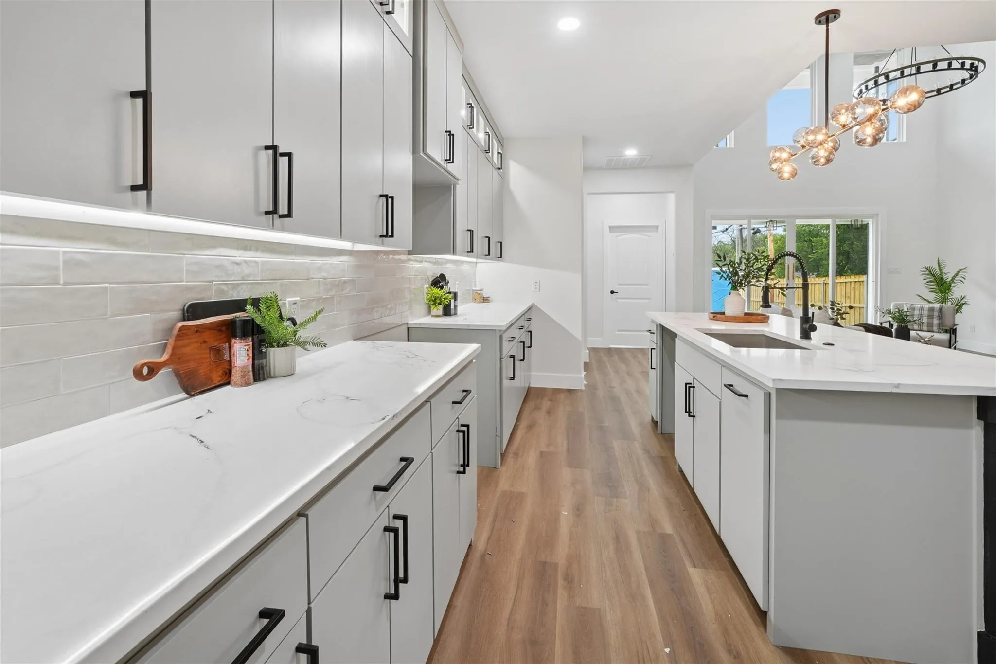 Modern galley kitchen featuring white cabinetry with black hardware, white quartz countertops, subway tile backsplash, stainless steel sink with faucet, wood-finish flooring, recessed and pendant lighting, and abundant natural light from oversized windows