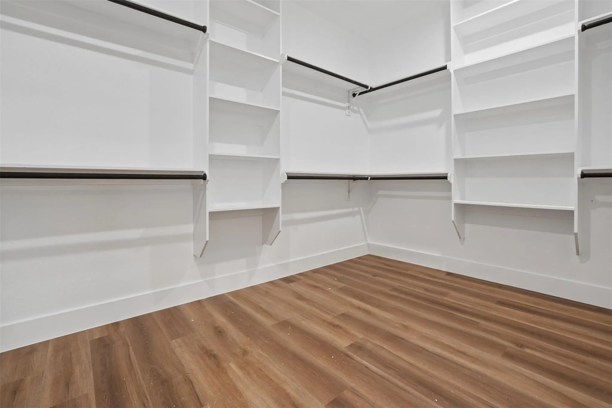 Spacious walk-in closet featuring custom white cabinetry with multiple shelving units, integrated hanging rods with metal hardware, and warm wood-finish flooring throughout