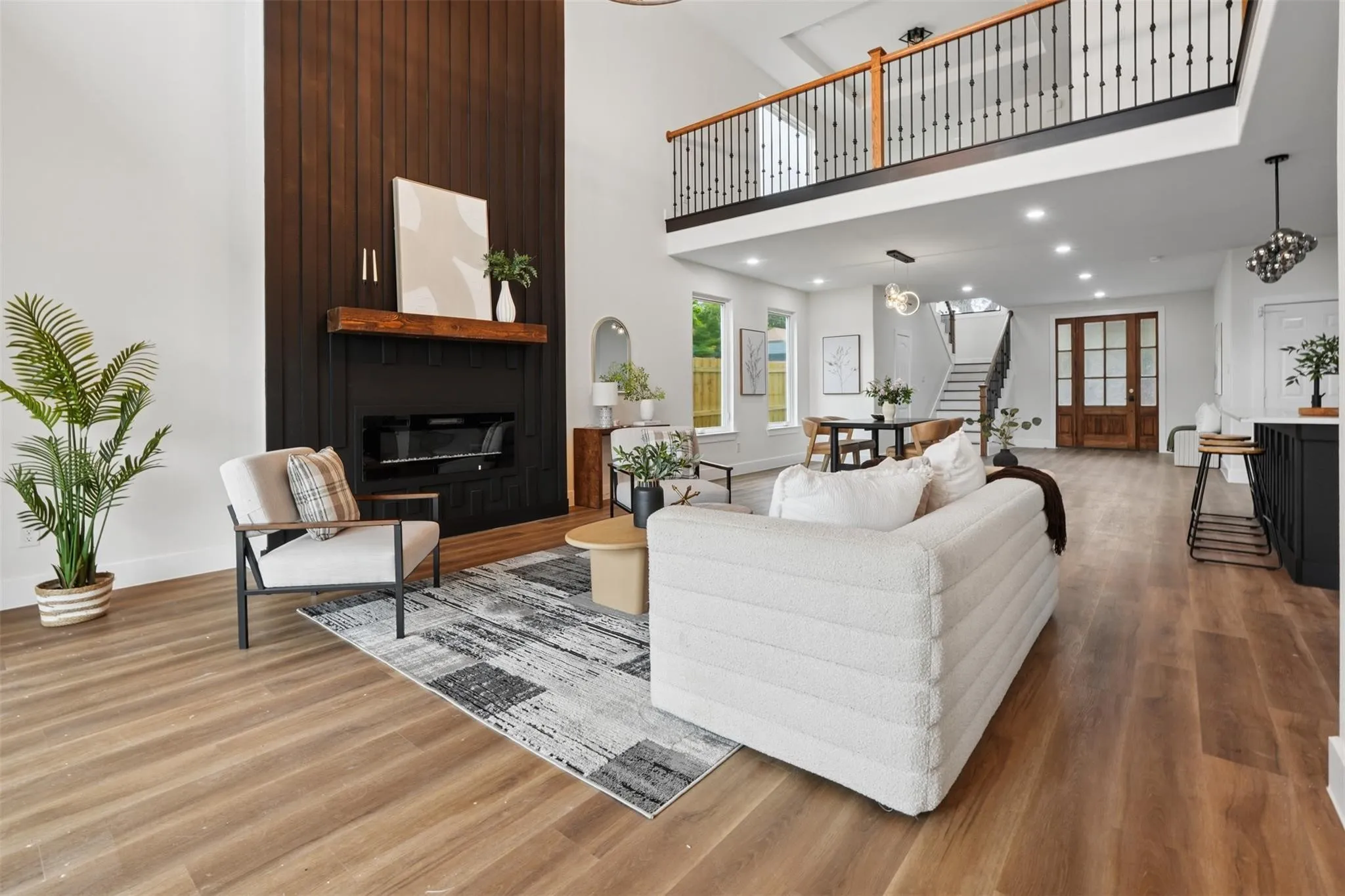 Two-story living area featuring a black paneled fireplace with a wood mantel, wood-finish flooring, and a second-story balcony with metal railings