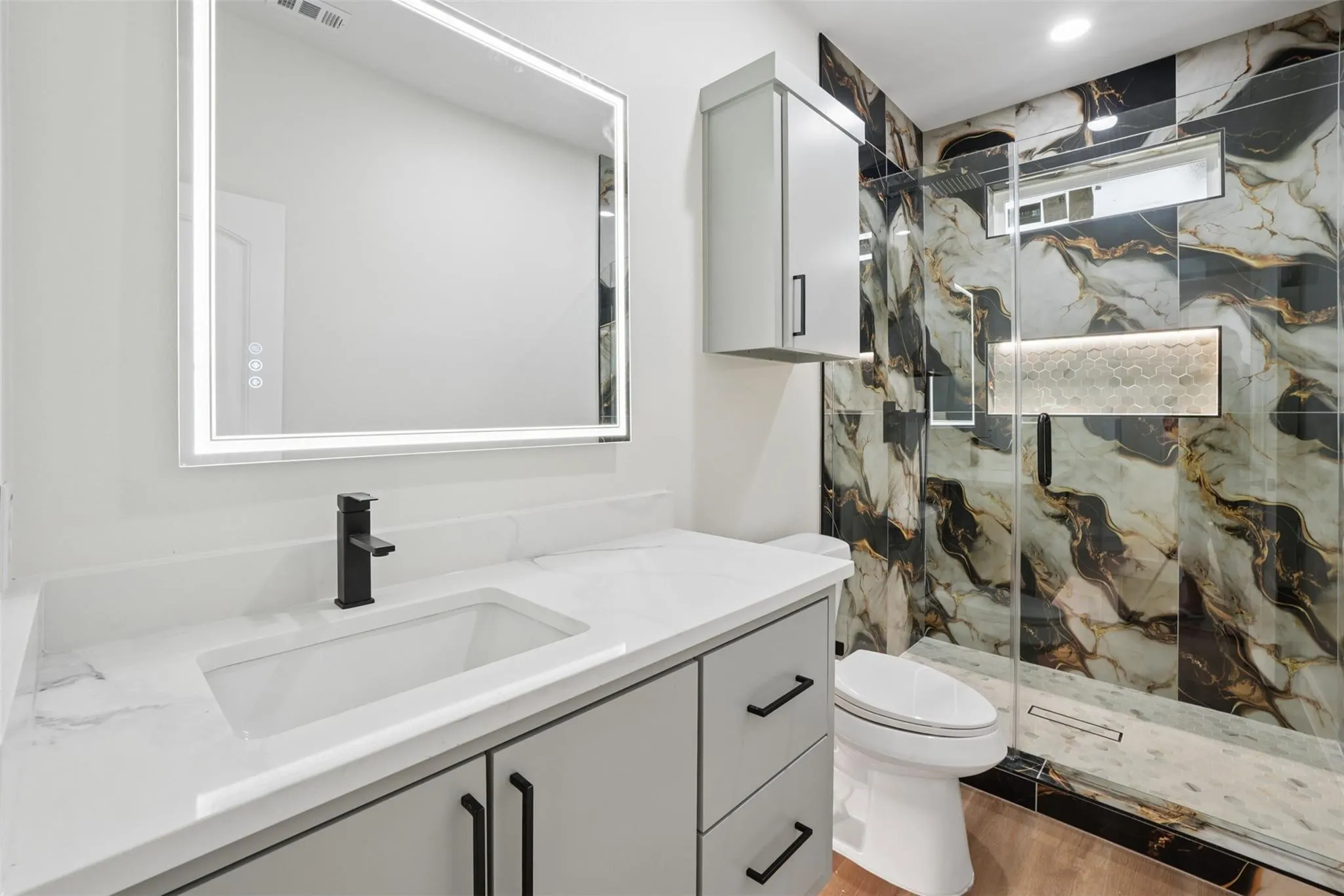 Bathroom featuring a large vanity with an integrated sink and quartz countertop, a backlit mirror, a glass-enclosed shower with decorative tile, and wood-finish flooring