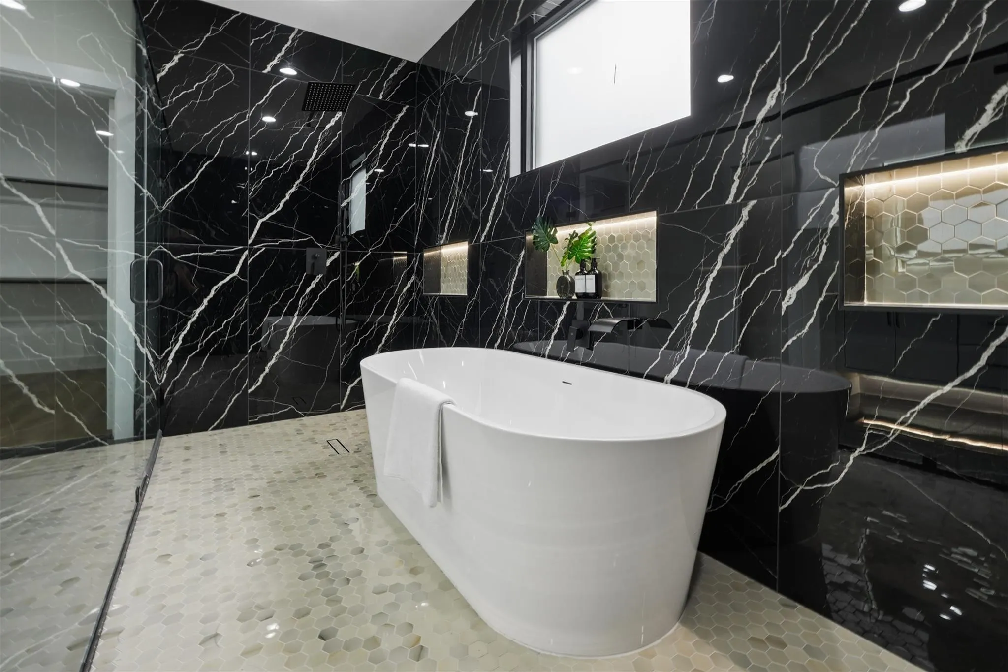 Freestanding soaking tub with floor-to-ceiling black marble-finish wall tiling, glass-enclosed shower, and illuminated recessed shelving