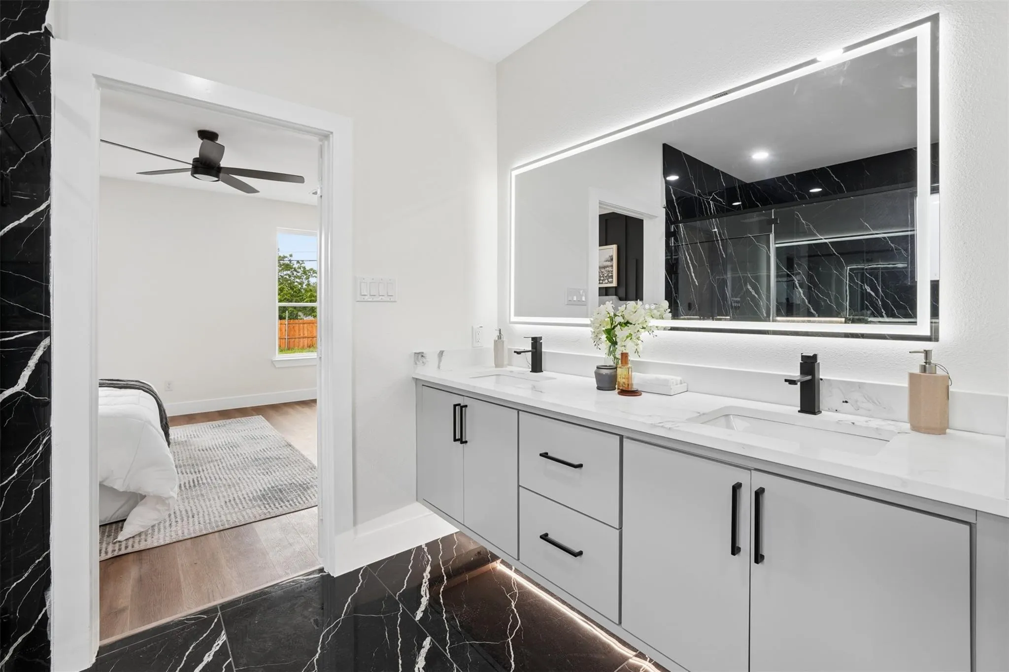 Dual sink vanity with matte black fixtures, an illuminated mirror, and a black marble-patterned accent wall