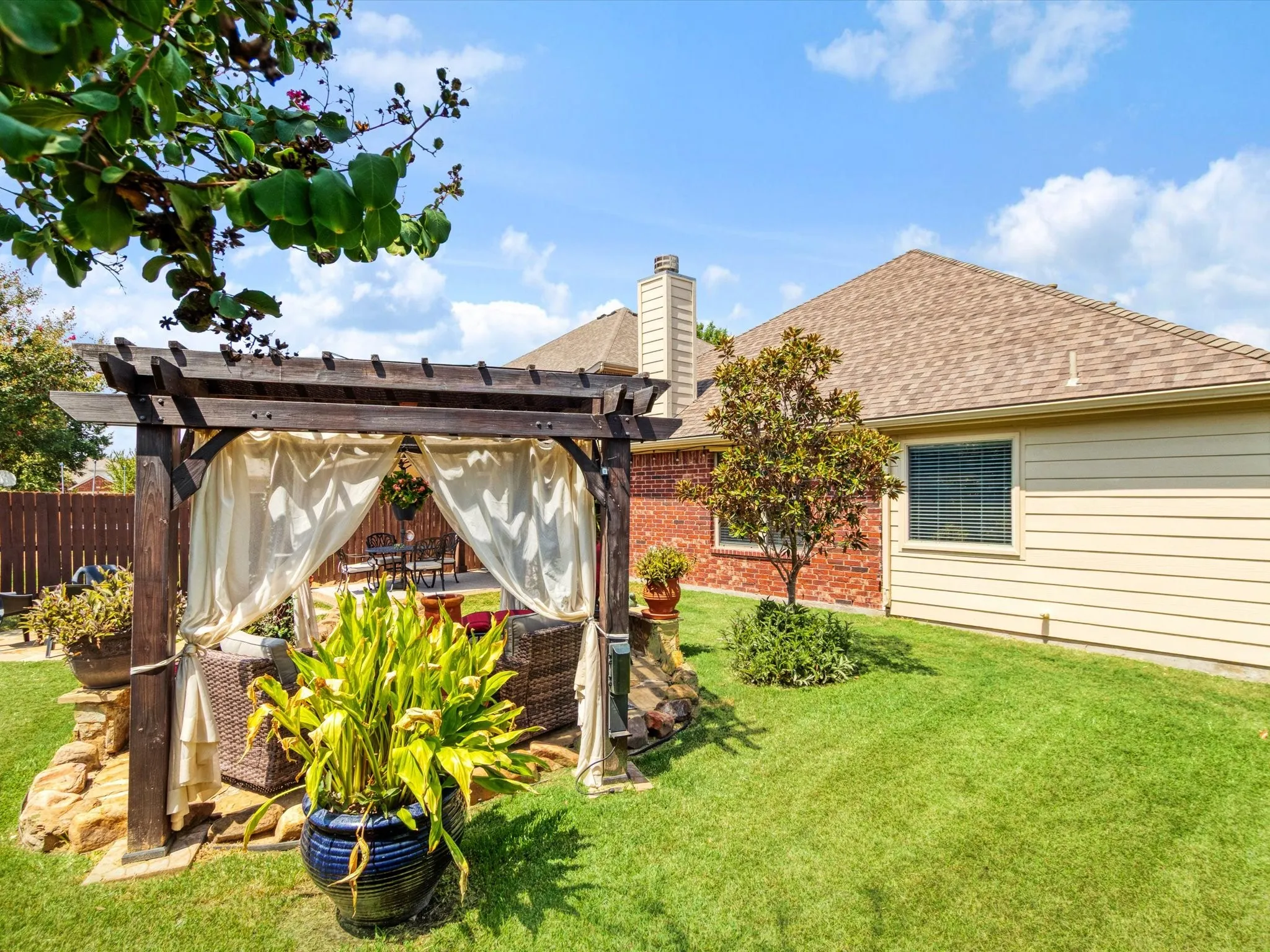 Expansive backyard featuring a manicured lawn, a wood pergola with draped fabric, and a stone-edged planting area