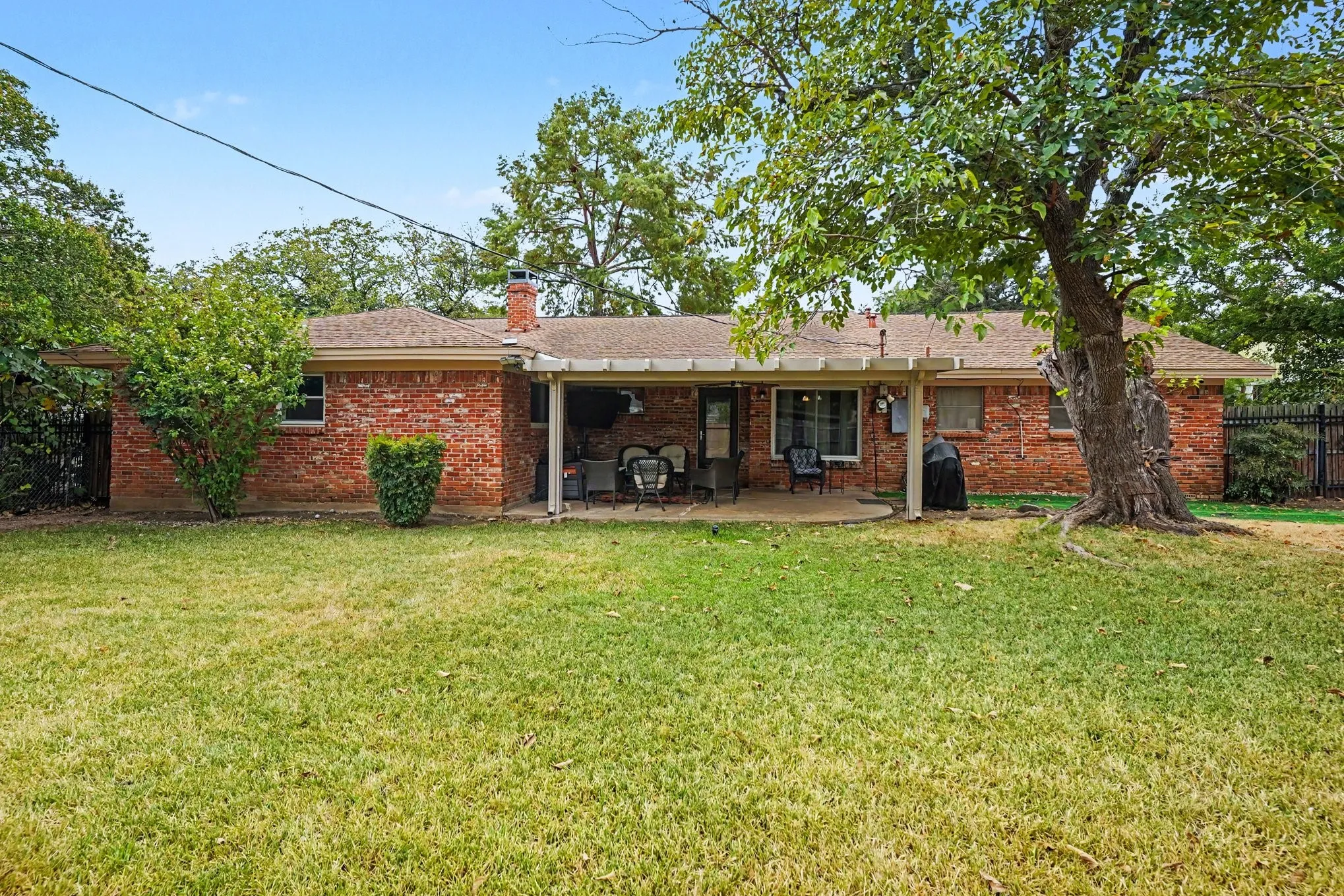Expansive backyard featuring a brick exterior, covered patio, mature trees, and a privacy fence