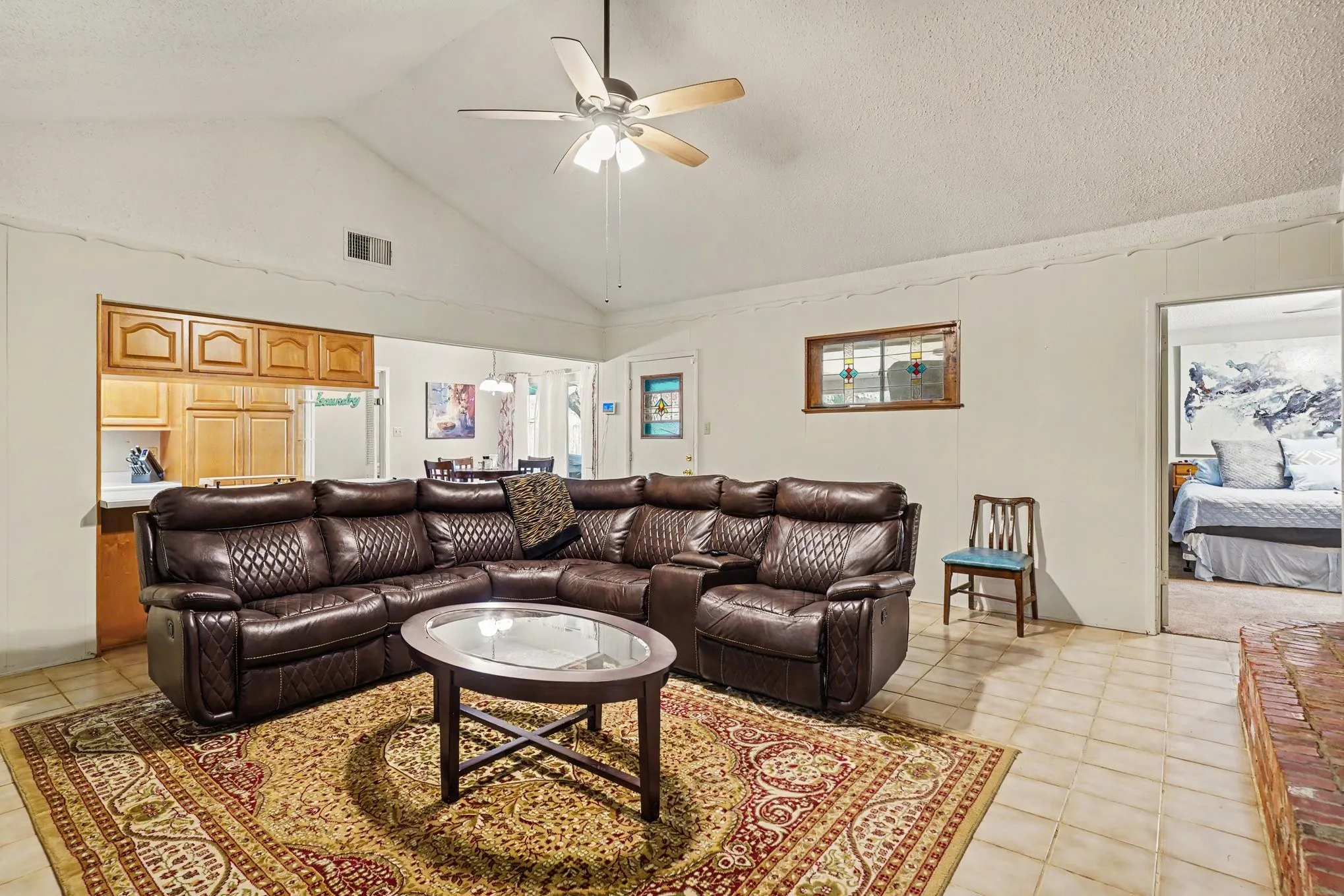 Vaulted ceiling with a ceiling fan, recessed kitchen pass-through, light-toned tile flooring, and a brick hearth