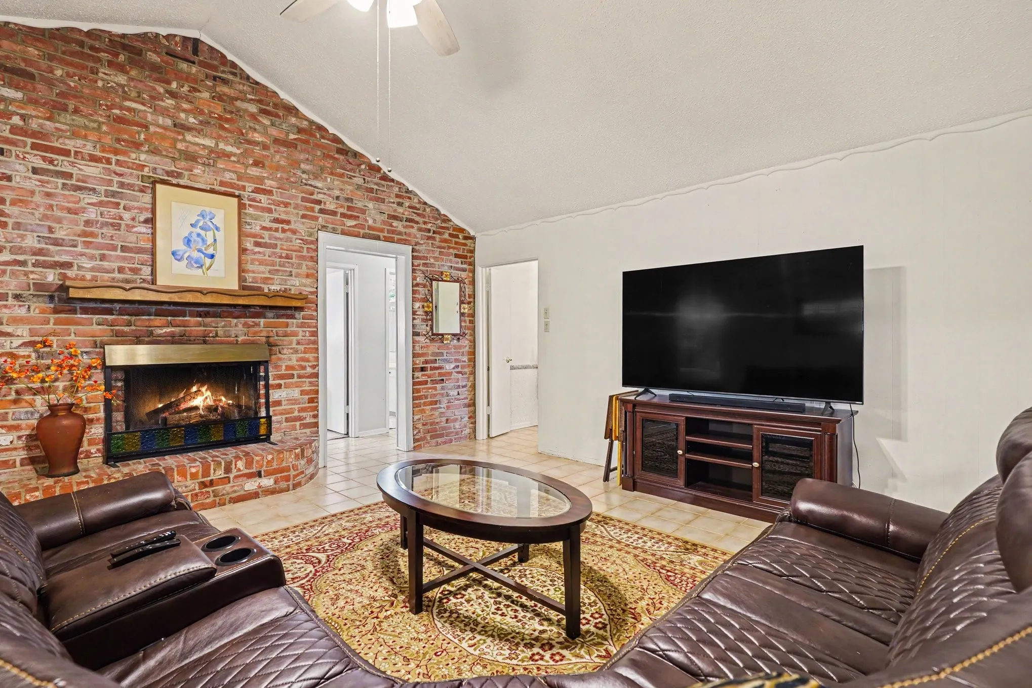 Vaulted ceiling, brick accent wall, built-in fireplace with mantel, and tile flooring