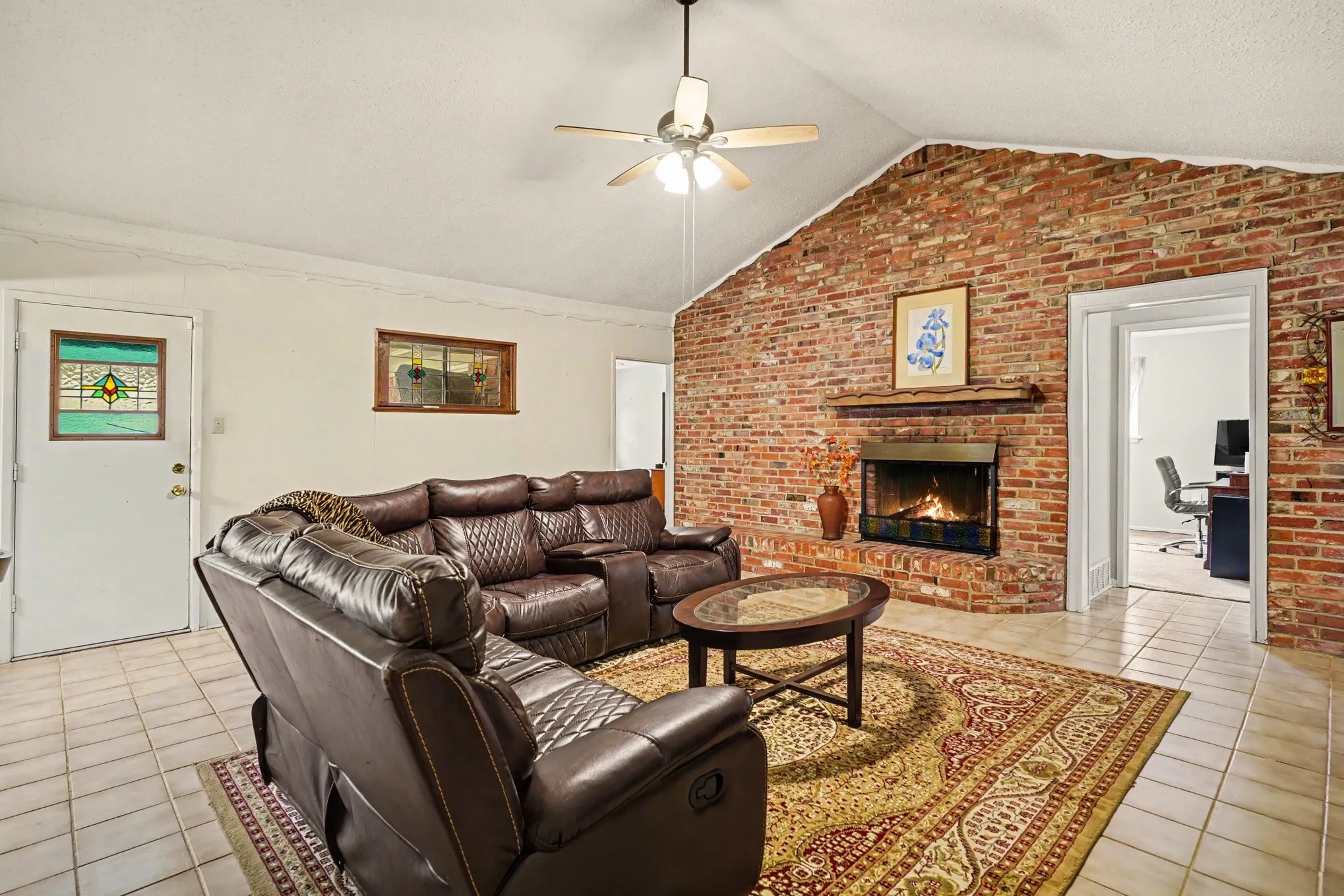Vaulted ceiling with a ceiling fan, exposed brick accent wall, wood-burning fireplace, and tiled flooring