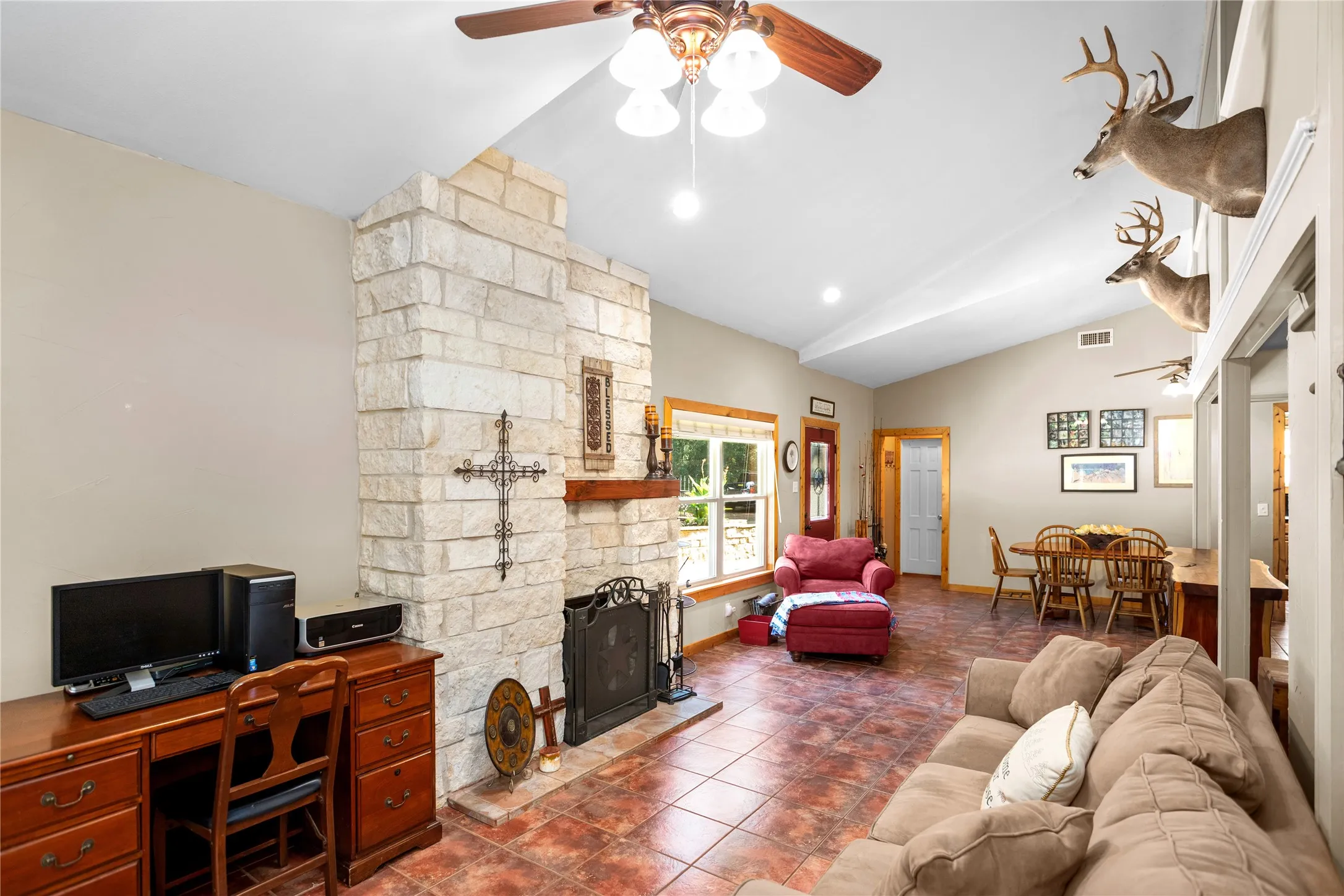Living room with ceiling fan, a stone fireplace, vaulted ceiling, tile flooring, and a desk