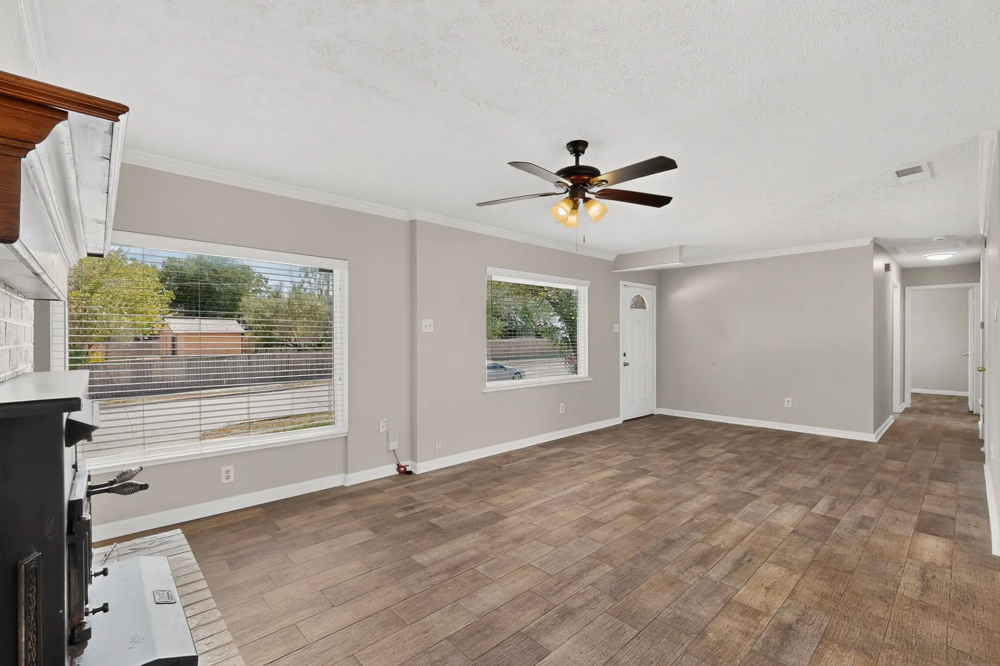 A bright living room featuring hardwood floors, a fireplace, a ceiling fan and wide windows, creating a spacious and airy atmosphere.