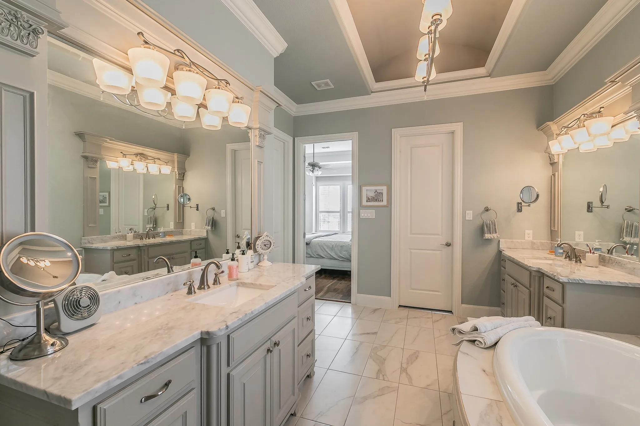 Dual vanities with beautiful countertops, tiled flooring, an oval soaking tub, and a decorative tray ceiling with an overhead light fixture