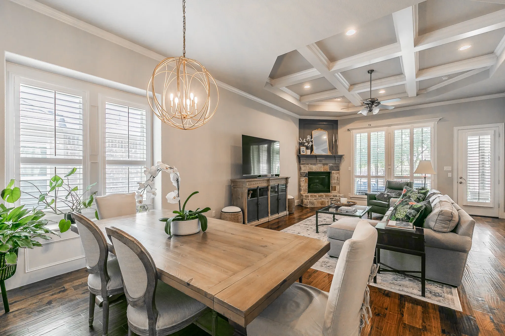 Rich wood-finish flooring throughout, featuring a stone-clad fireplace, coffered ceiling, and plantation shutters
