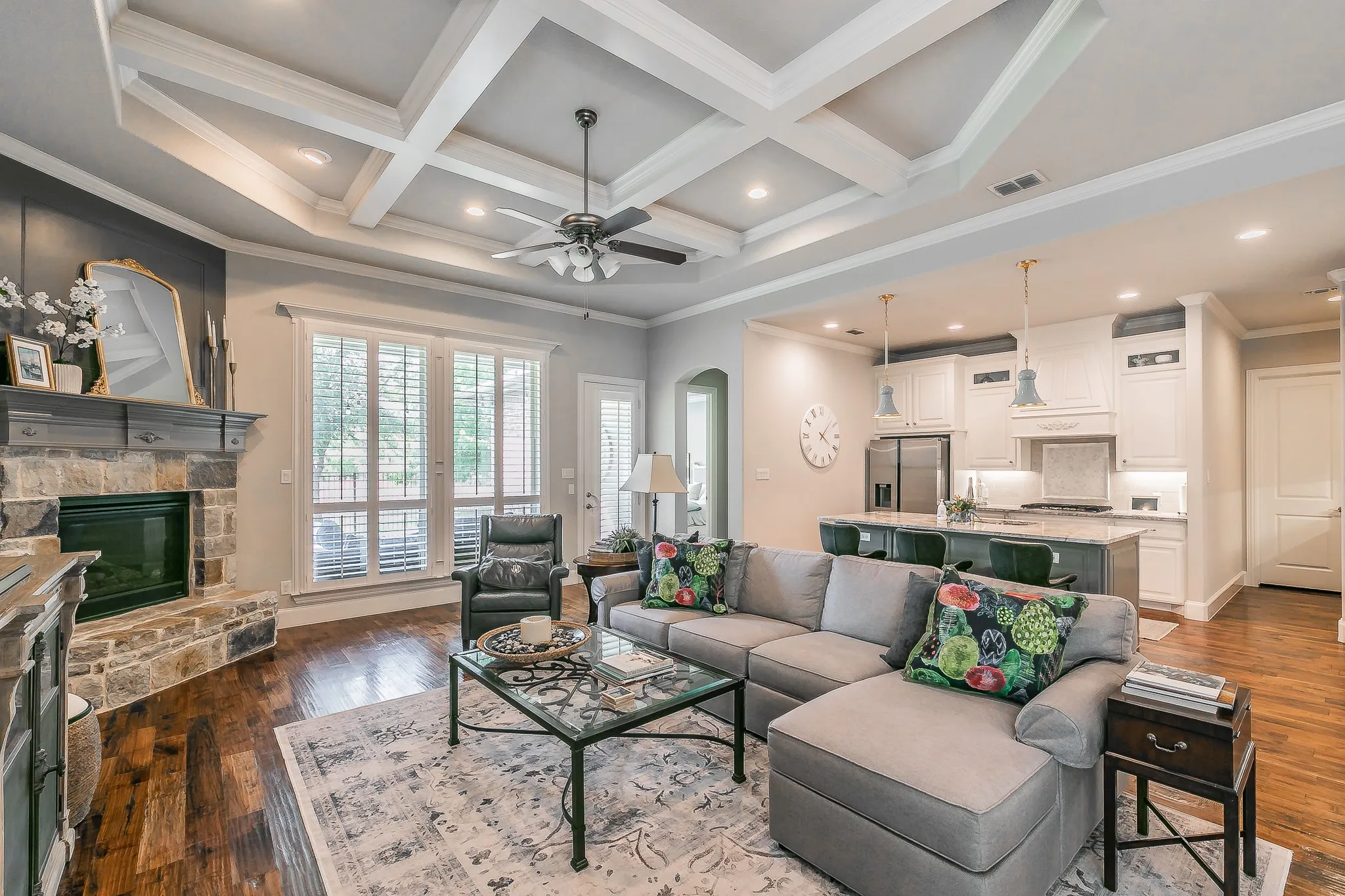 Coffered ceilings, stone-clad fireplace with mantel,  wood flooring, plantation shutters, and a kitchen featuring white cabinetry and a central island with pendant lighting