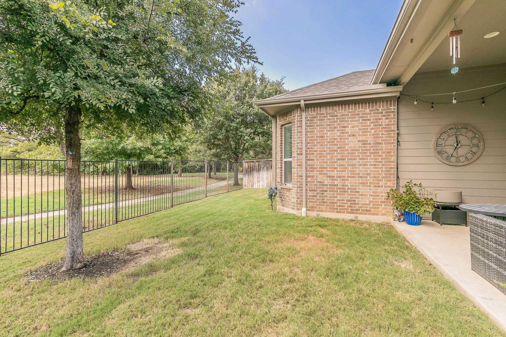 Brick exterior with a covered patio, string lighting, and an adjacent grassy area with mature trees and a black metal fence