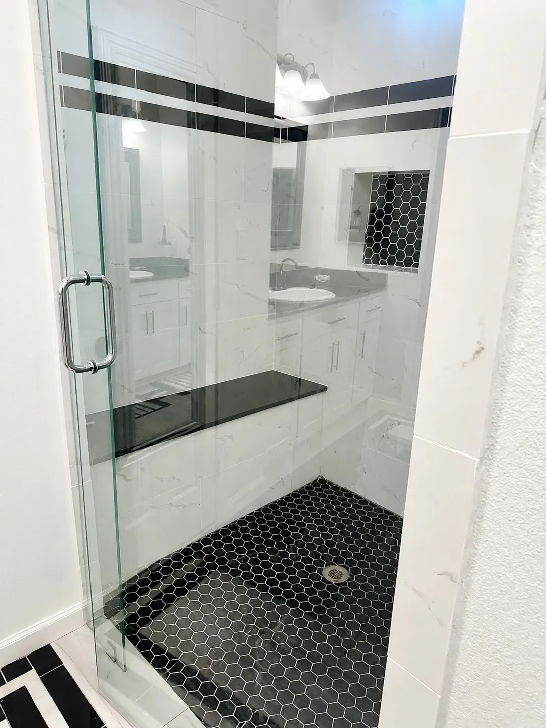 Glass-enclosed shower with black hexagonal floor tile, white marble-patterned wall tile, a black contrasting accent stripe, a built-in black bench, and a tiled shower niche