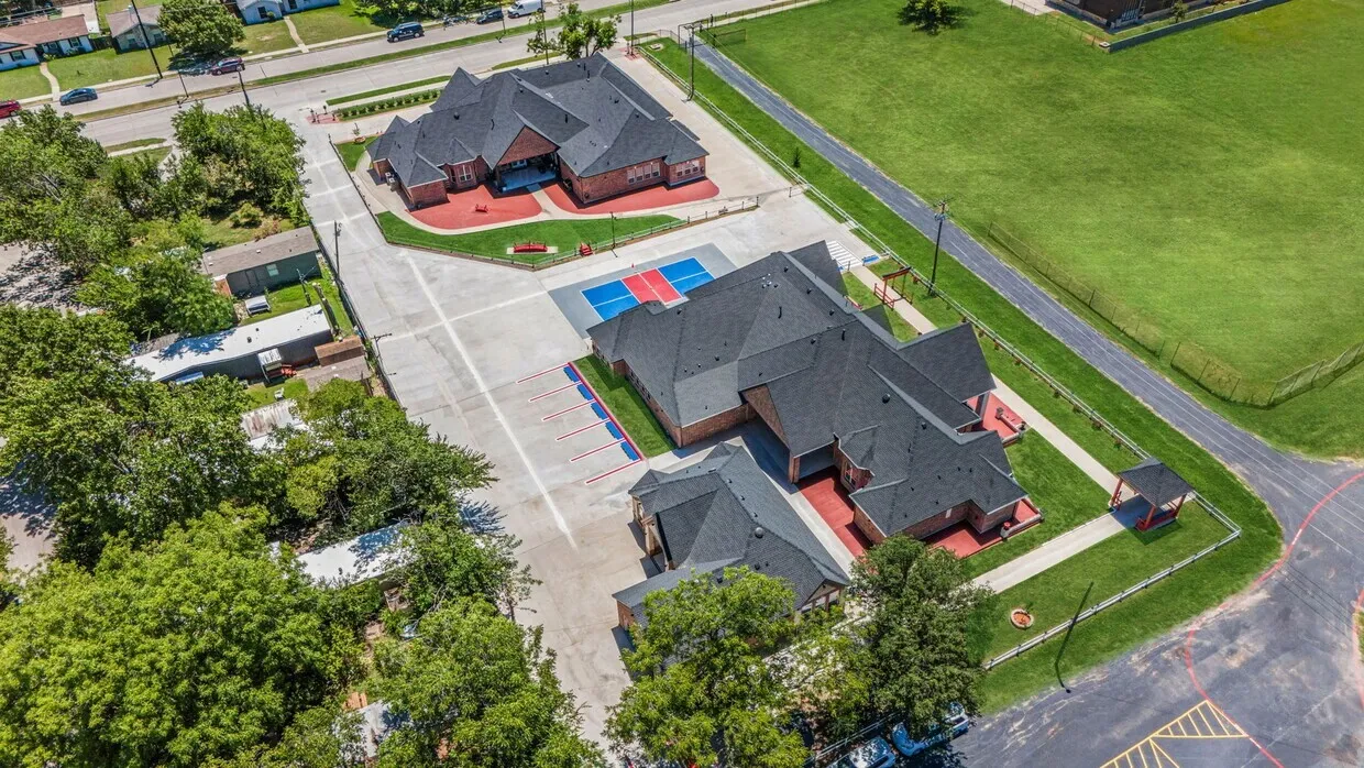Expansive paved parking area, two brick structures with dark shingle roofs, dedicated athletic court, and a gazebo on a manicured lawn