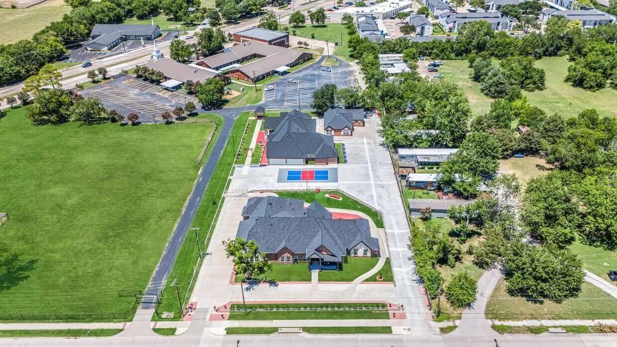 Expansive property featuring multiple buildings with dark shingle roofs, substantial paved hardscaping, and distinct green spaces