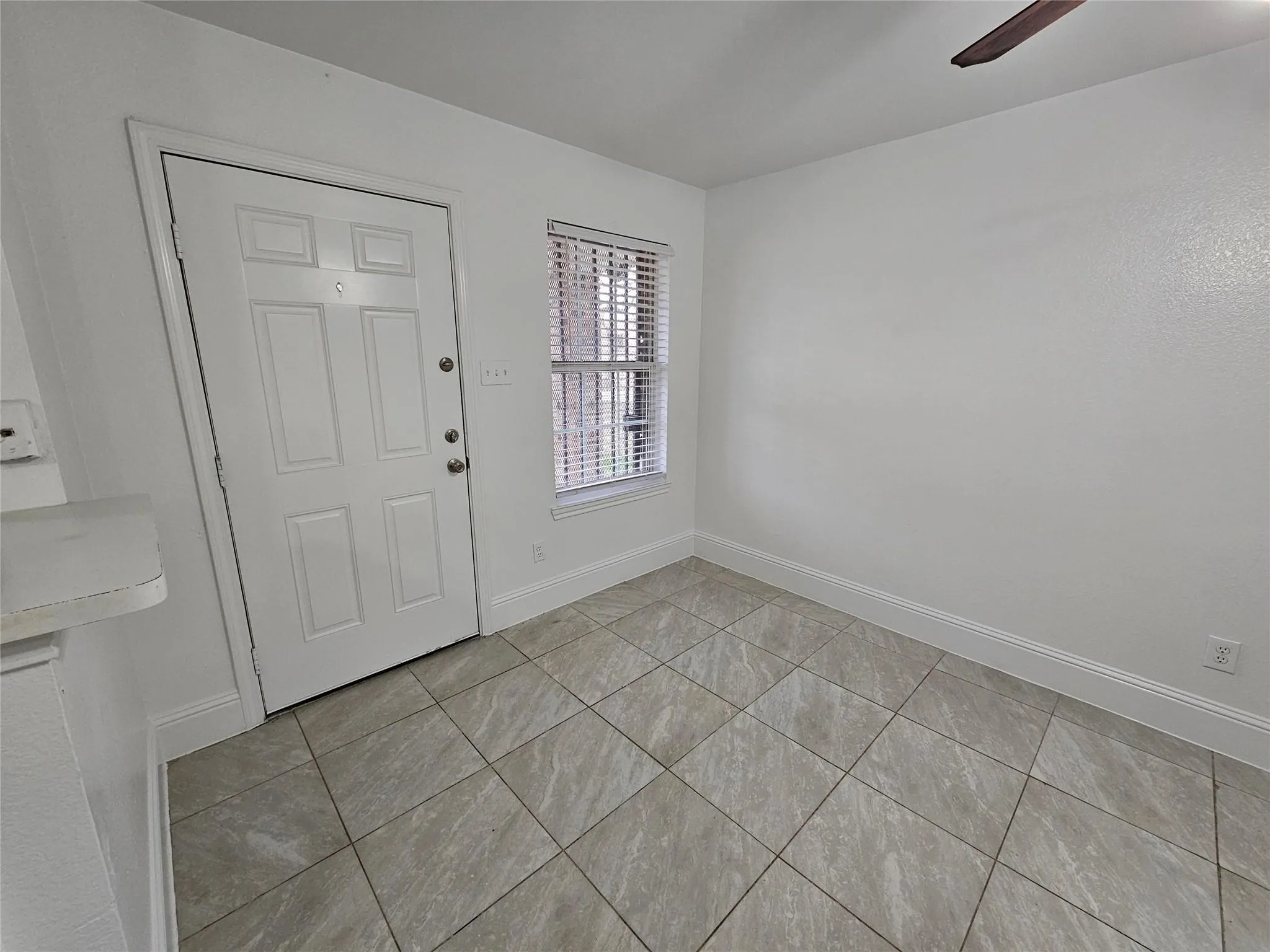 White paneled door, window with blinds, wood-finish ceiling fan, neutral tile flooring, and white baseboards