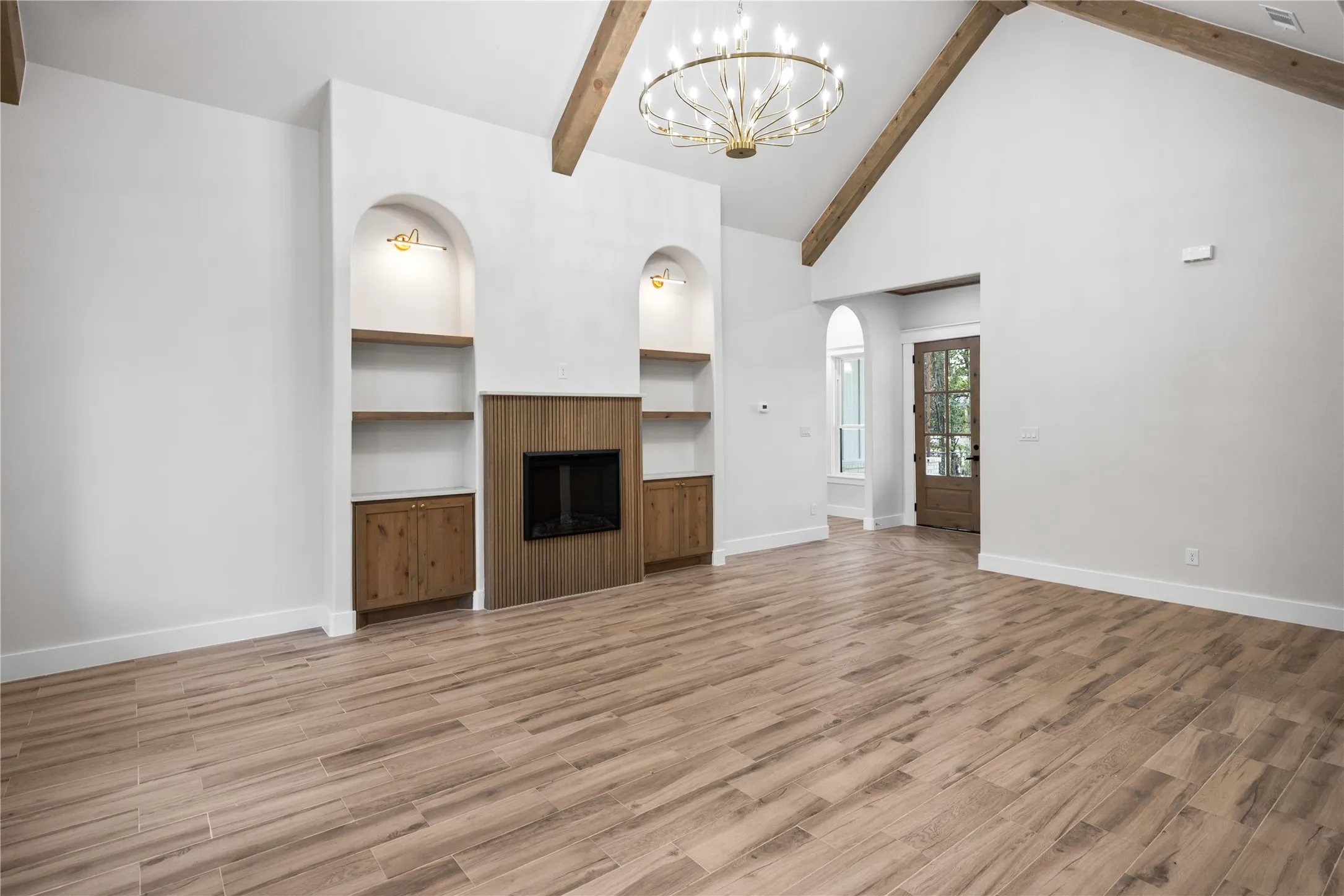 Vaulted ceiling with exposed wood beams, wood-finish flooring, built-in shelving with integrated lighting, wood-paneled fireplace surround, and a decorative chandelier