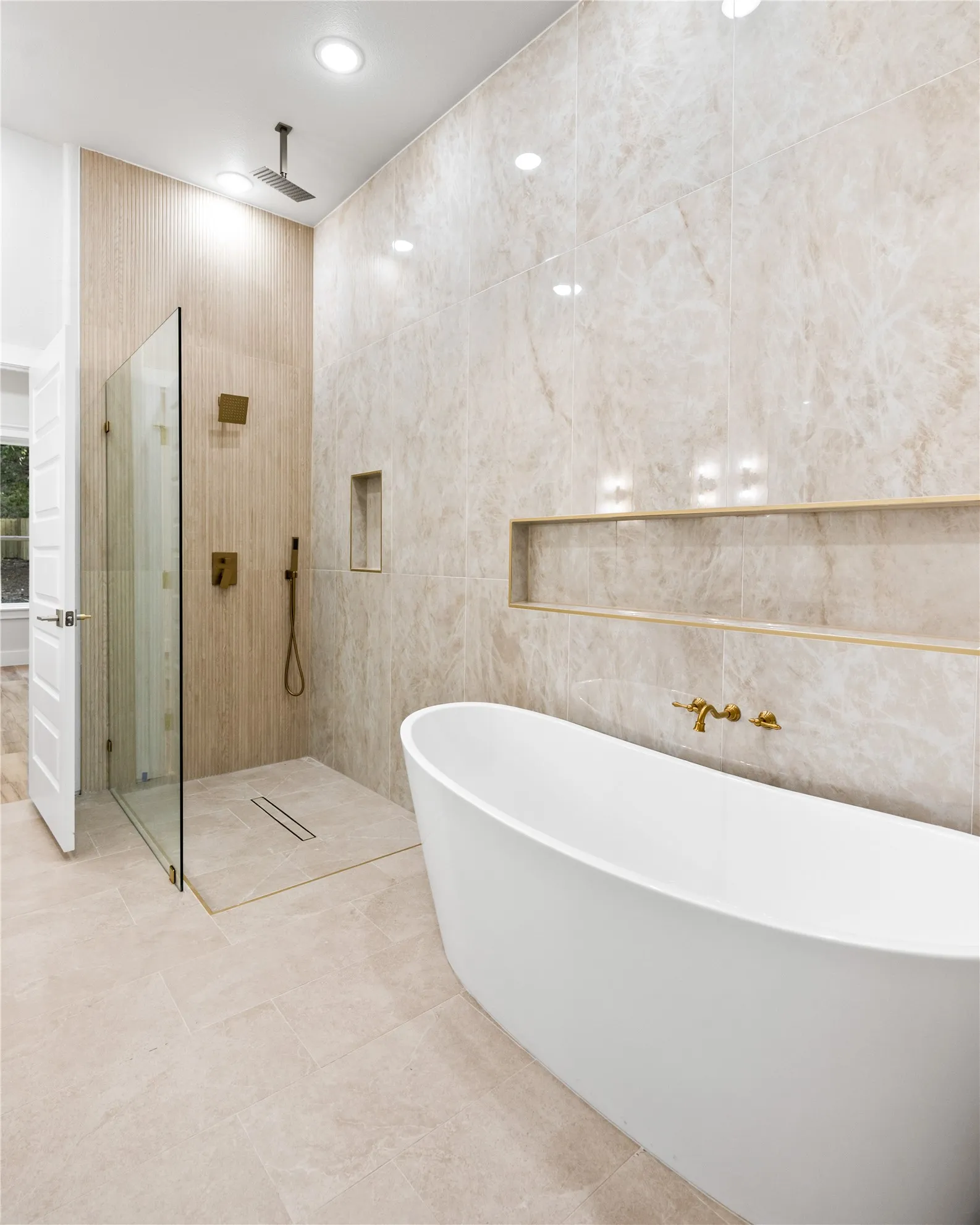 Freestanding soaking tub, walk-in shower with glass enclosure, wall-mounted shower fixtures, recessed shower niche, and large format floor and wall tiling