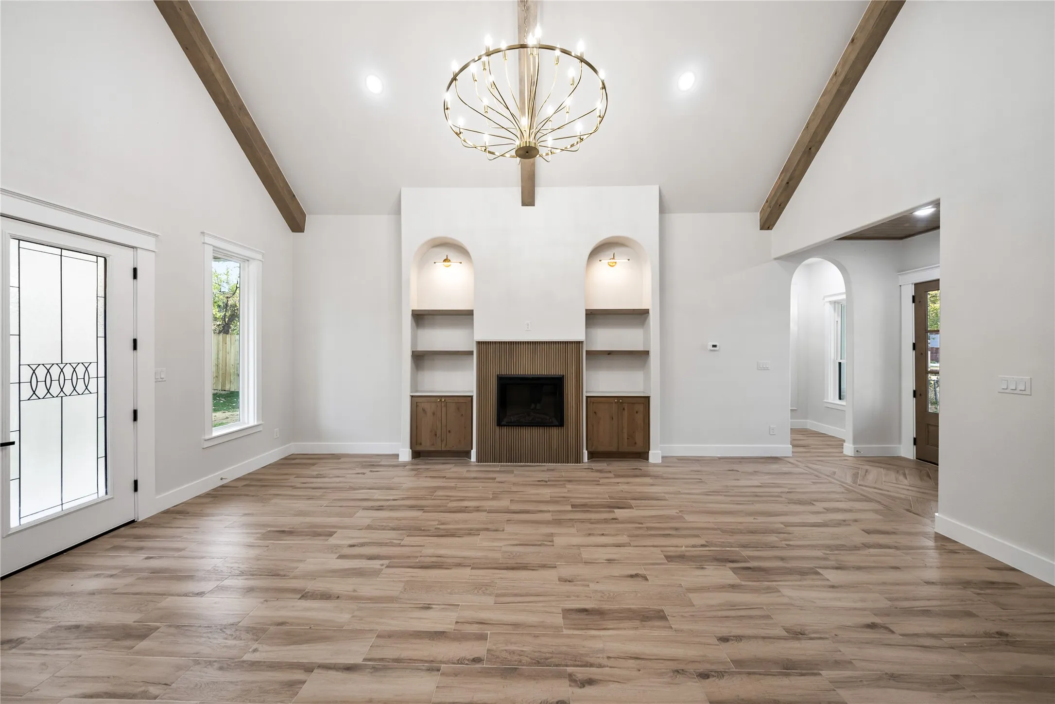 Vaulted ceiling with exposed wood beams, wood-finish flooring, central fireplace with built-in cabinetry, decorative arched niches, and an ornate chandelier