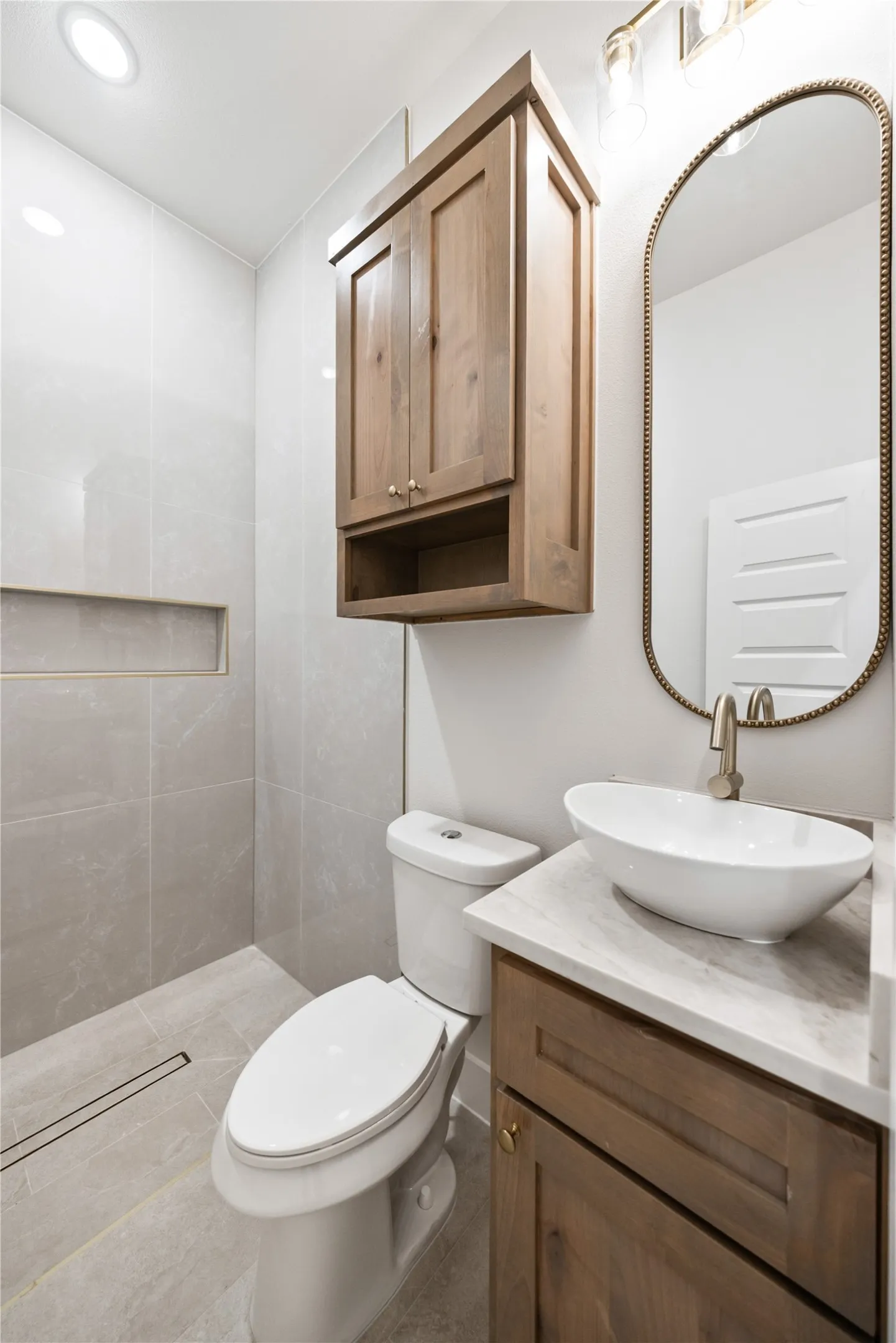Vessel sink, wood-finish vanity with gold-tone hardware, wall-mounted wood-finish cabinet, and a tiled shower with a linear drain