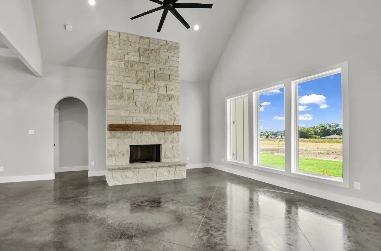 Vaulted ceiling with a ceiling fan, stone fireplace with a wood mantle, three vertical windows, archway, and polished concrete flooring