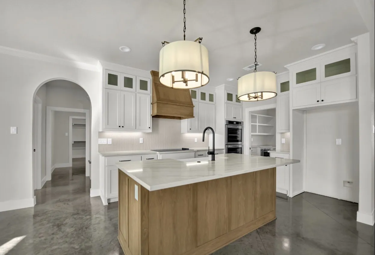 Gourmet kitchen featuring a wood-finish island with waterfall countertop edge, white shaker cabinetry with glass-front upper doors, polished concrete flooring, stainless steel appliances, pendant lighting, and a waterfall-edge quartz countertop at the sink station
