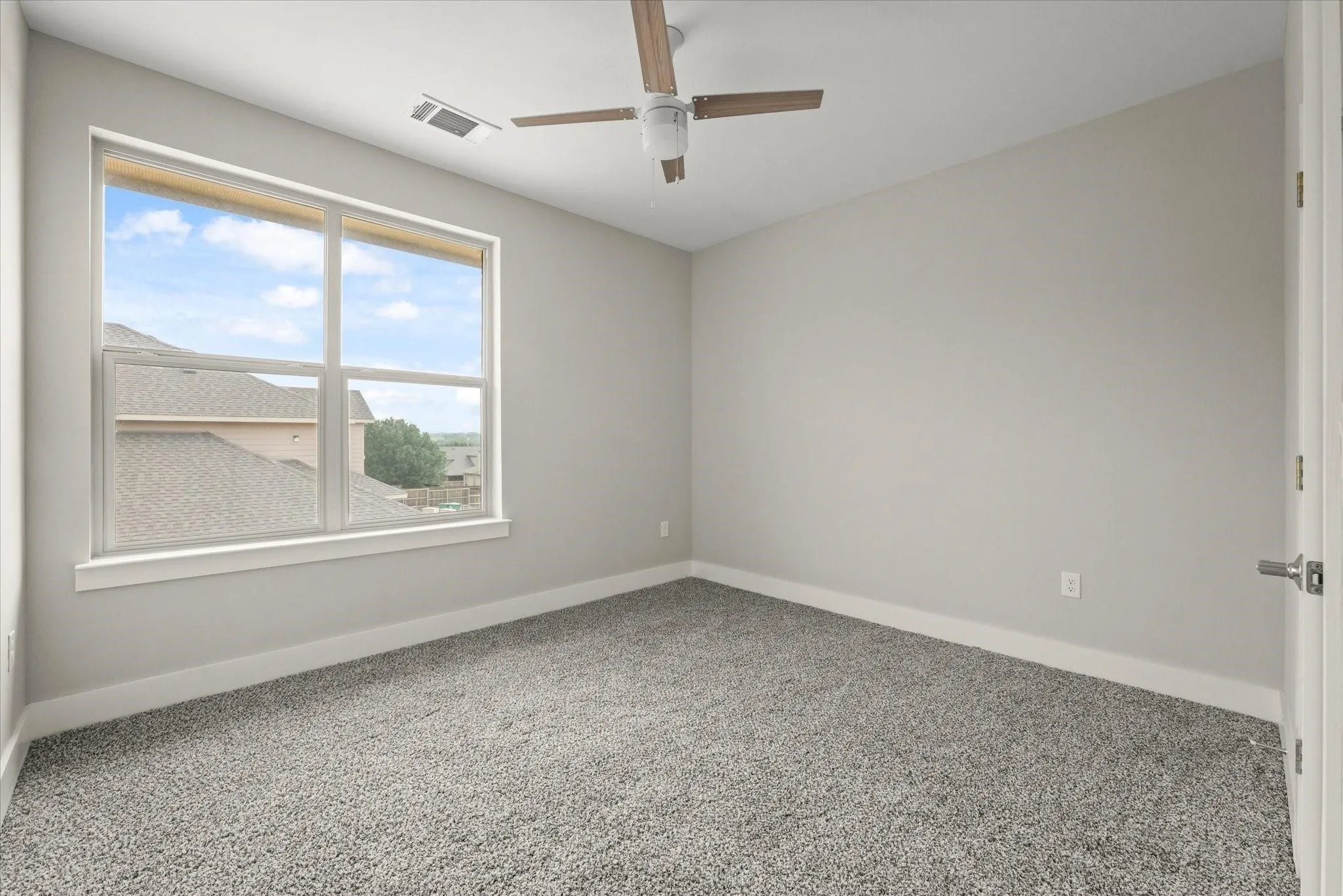 Bright room featuring a large window, plush gray carpeting, neutral wall tones, and a ceiling fan