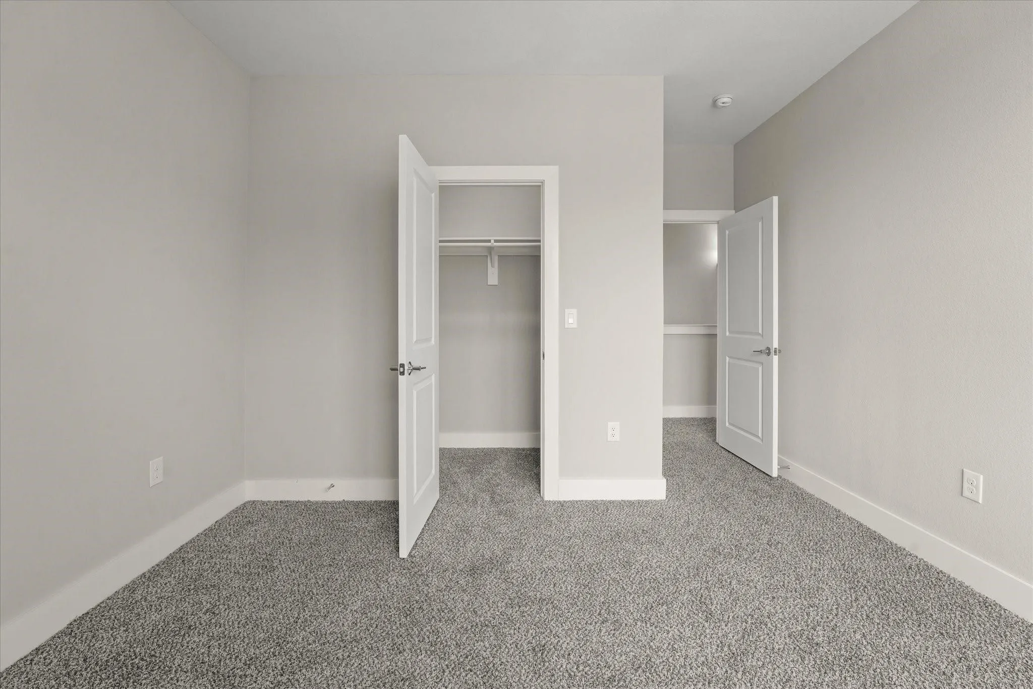 Neutral-toned walls with grey carpeting, featuring two closets with white bi-fold doors and integrated shelving
