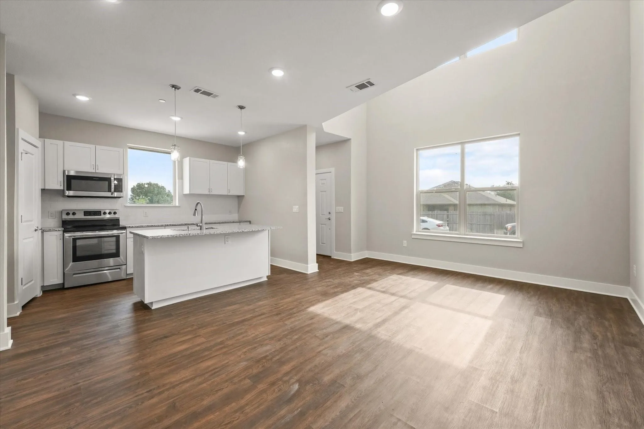 Vaulted ceiling with high windows, wood-finish flooring, white cabinetry, stainless steel appliances, and a kitchen island with pendant lighting