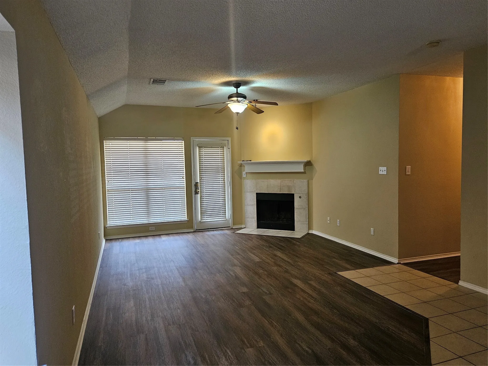 Vaulted ceiling, wood-finish flooring, corner fireplace with tile surround, ceiling fan with light fixture, and large window with blinds