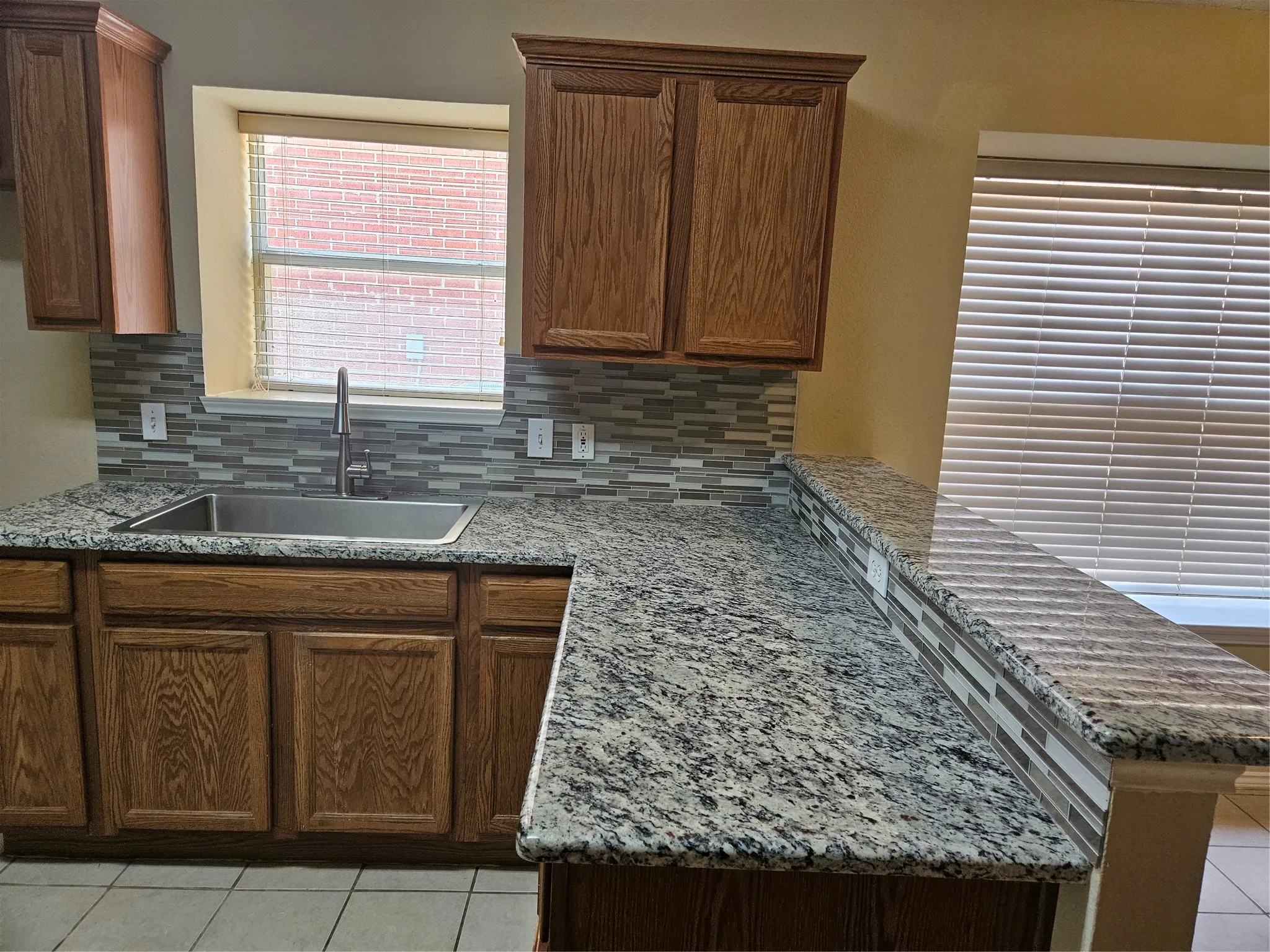 Granite countertops with an integrated stainless steel sink, wood cabinetry, and a mosaic tile backsplash