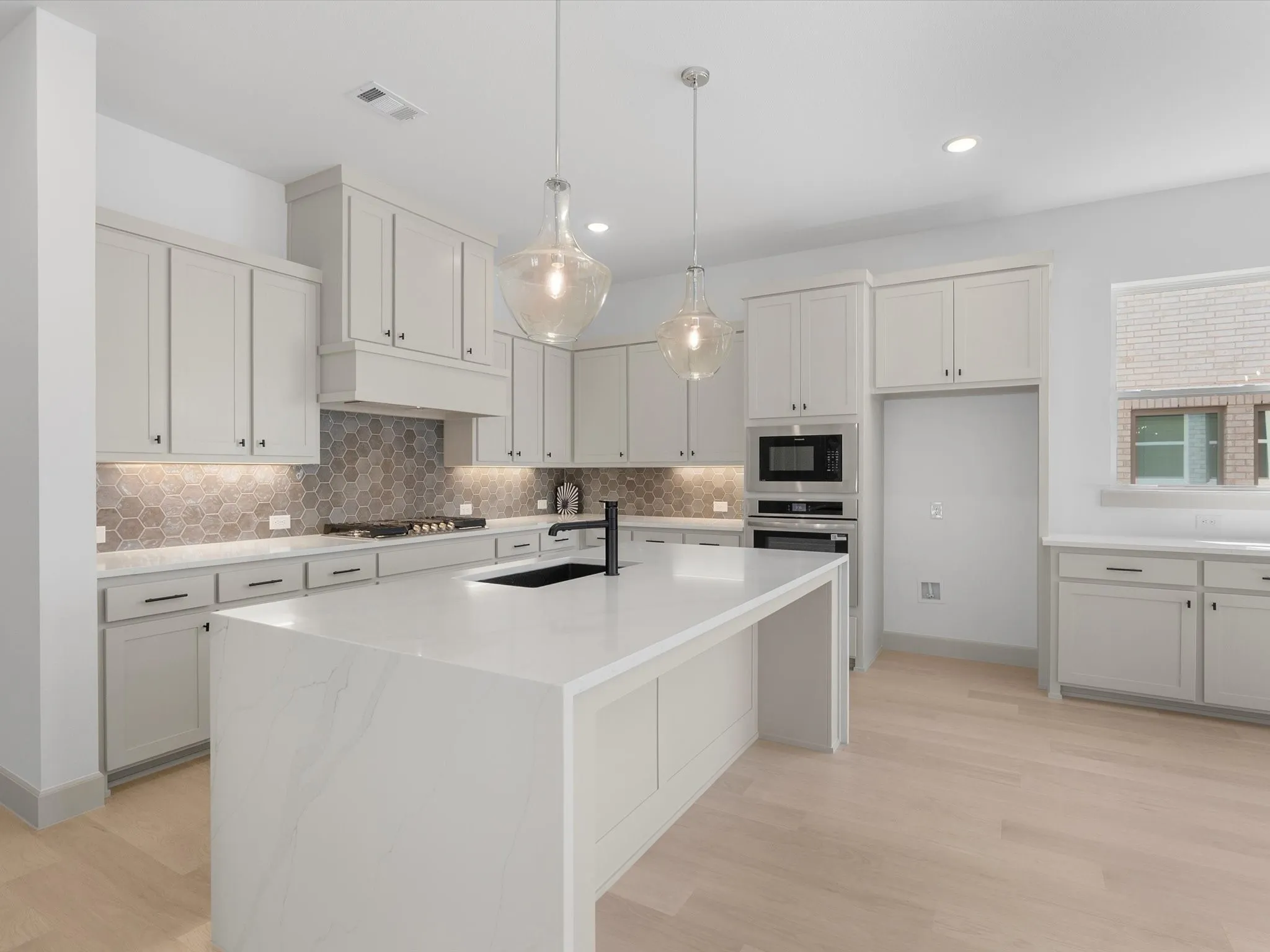 Bright kitchen with light-toned wood-finish flooring, white cabinetry, a built-in oven and microwave, a large island with an integrated sink, and pendant lighting