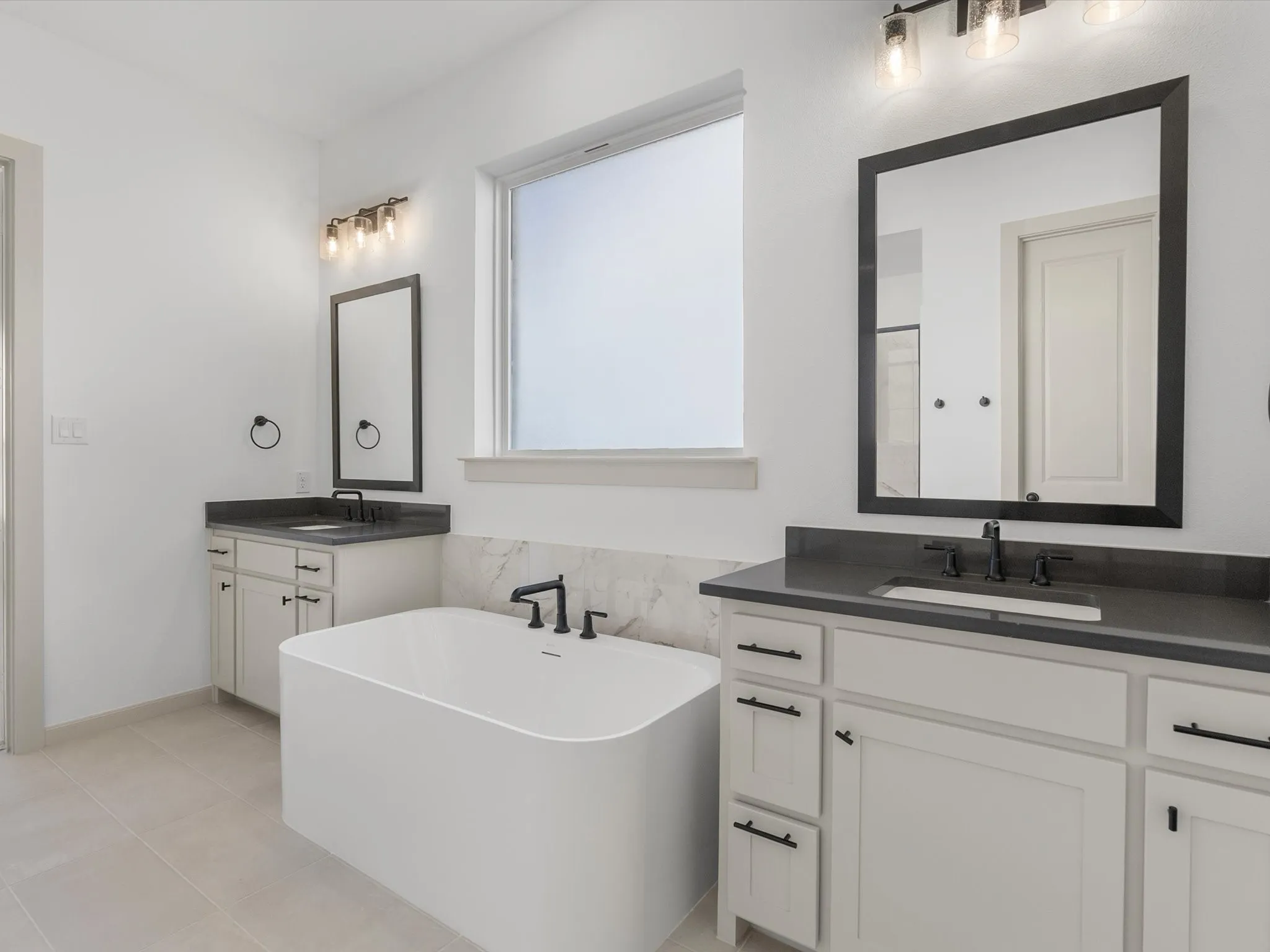 Freestanding soaking tub with a wall-mounted faucet, dual vanities with dark countertops, and light-toned tile flooring