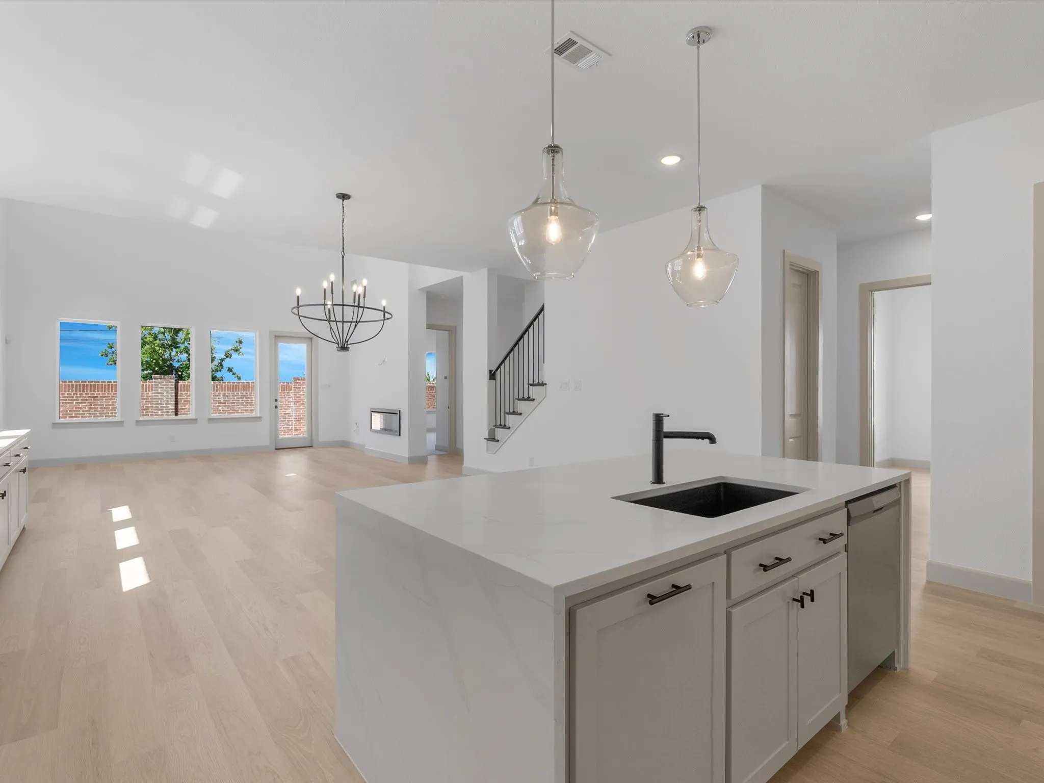 Open-concept living space with light wood-finish flooring, a kitchen island featuring a black sink and faucet, pendant lighting, and a modern chandelier