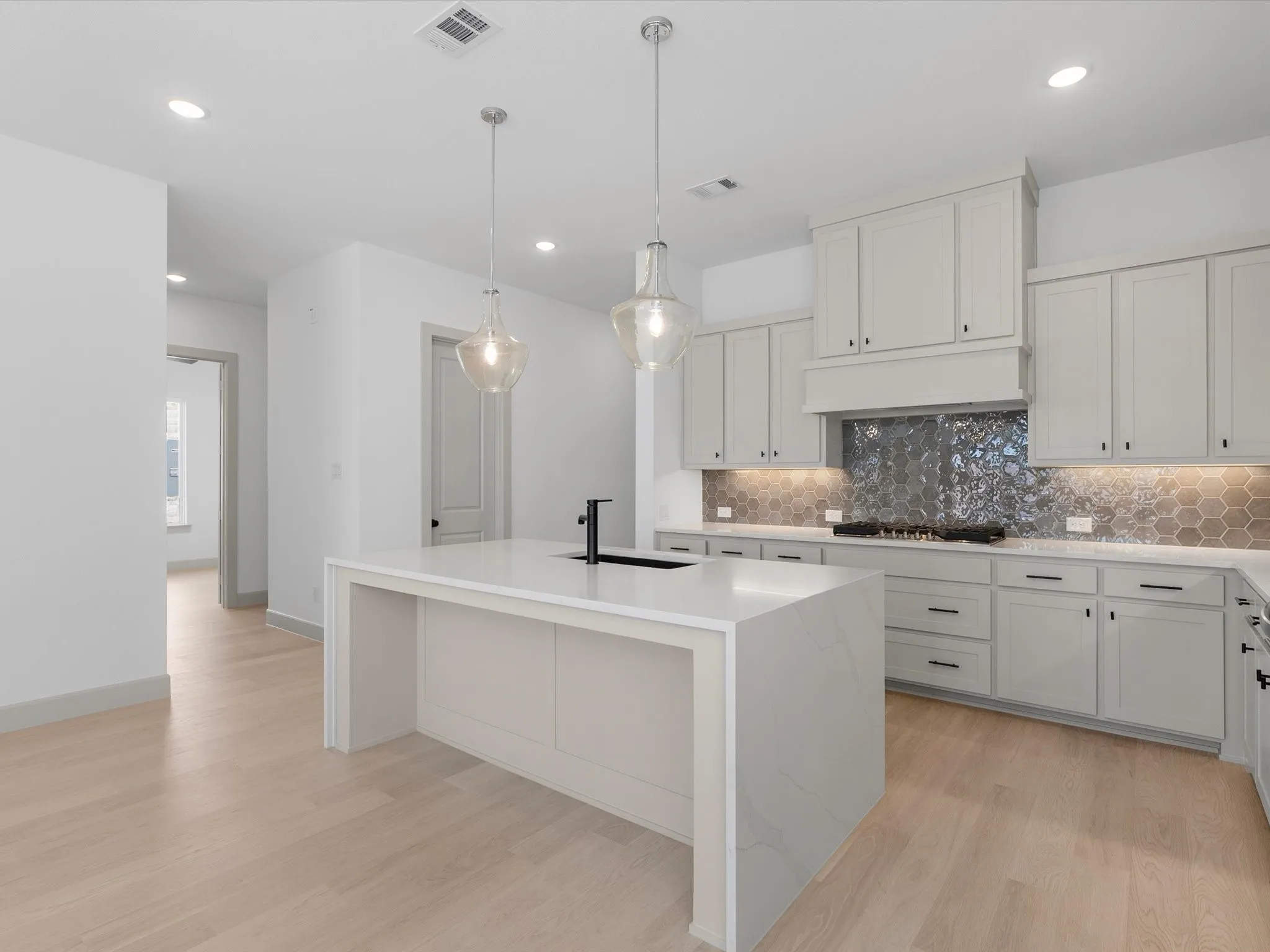 Kitchen featuring a large waterfall island with a built-in sink, pendant lighting, light-toned cabinetry, quartz countertops, and a hexagonal tile backsplash