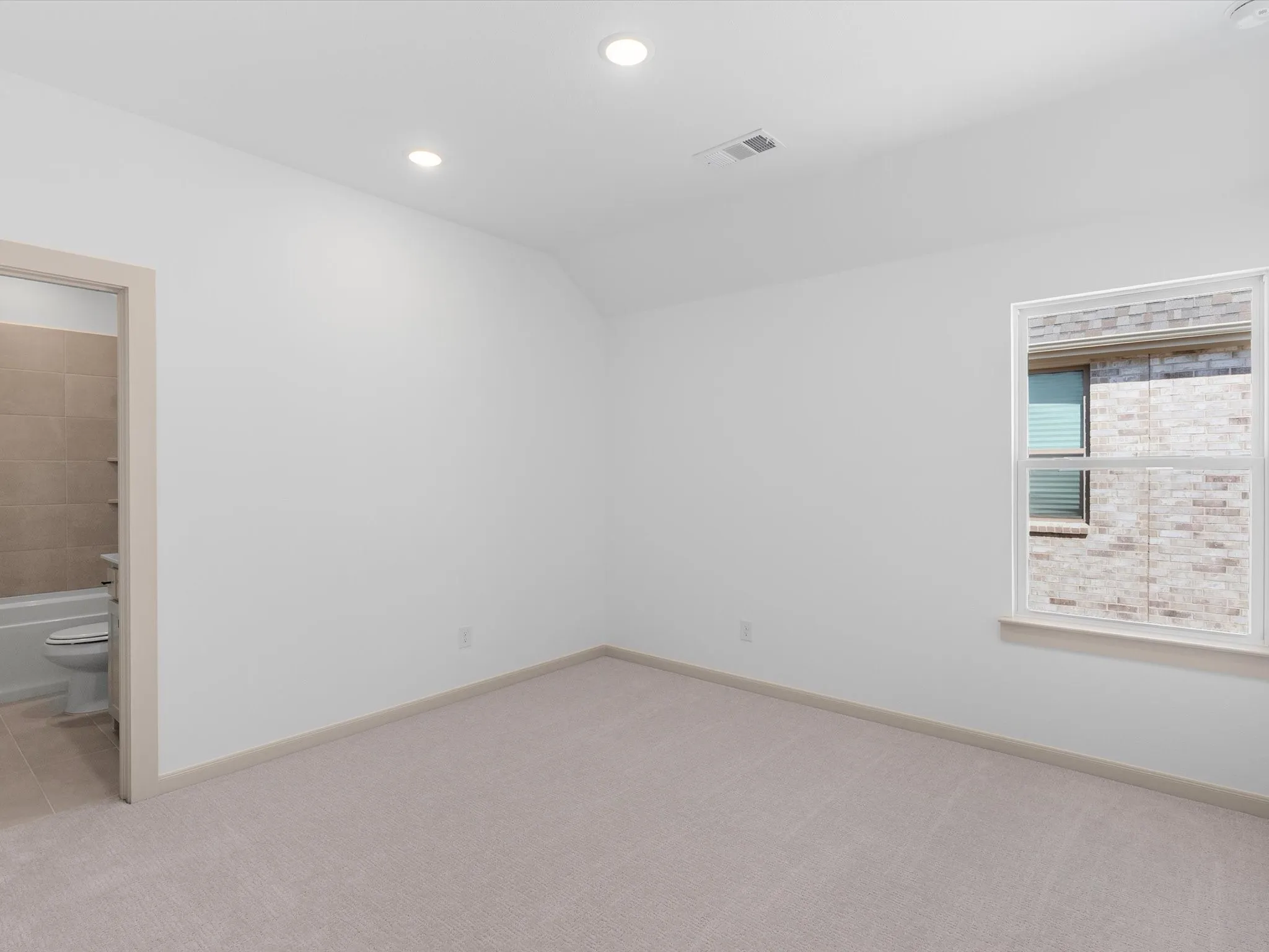 Recessed ceiling lighting, vaulted ceiling, neutral tone carpet flooring, window with exterior brick detail, and attached bathroom with tiled shower surround