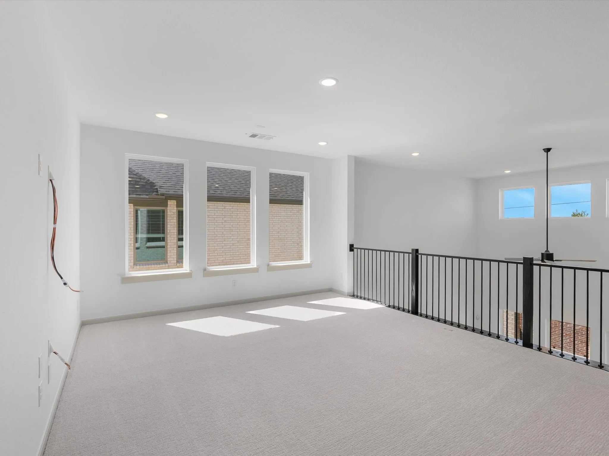 Spacious loft area featuring light-colored carpeting, multiple windows providing ample natural light, recessed ceiling lighting, a contemporary ceiling fan, and a black wrought iron railing