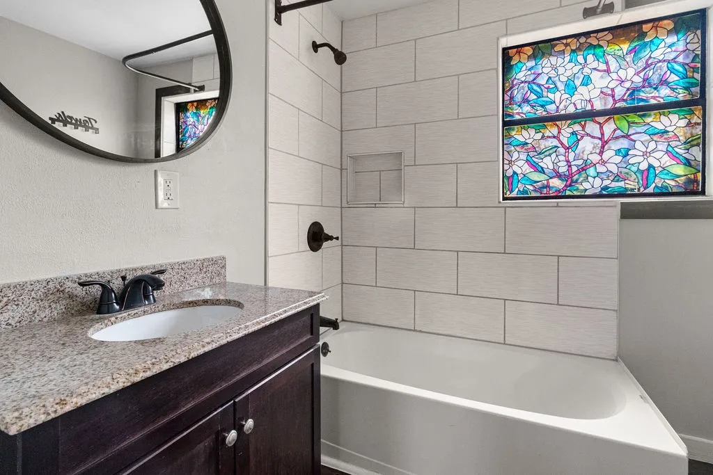 Built-in bathtub with subway tile surround, integrated shower niche, stained glass window, vanity with granite countertop, and oval undermount sink