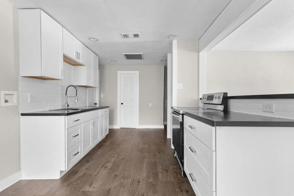 Modern galley kitchen featuring white cabinetry with brushed metal hardware, black countertops, stainless steel appliances, a chrome pull-down faucet, and wood-finish flooring throughout