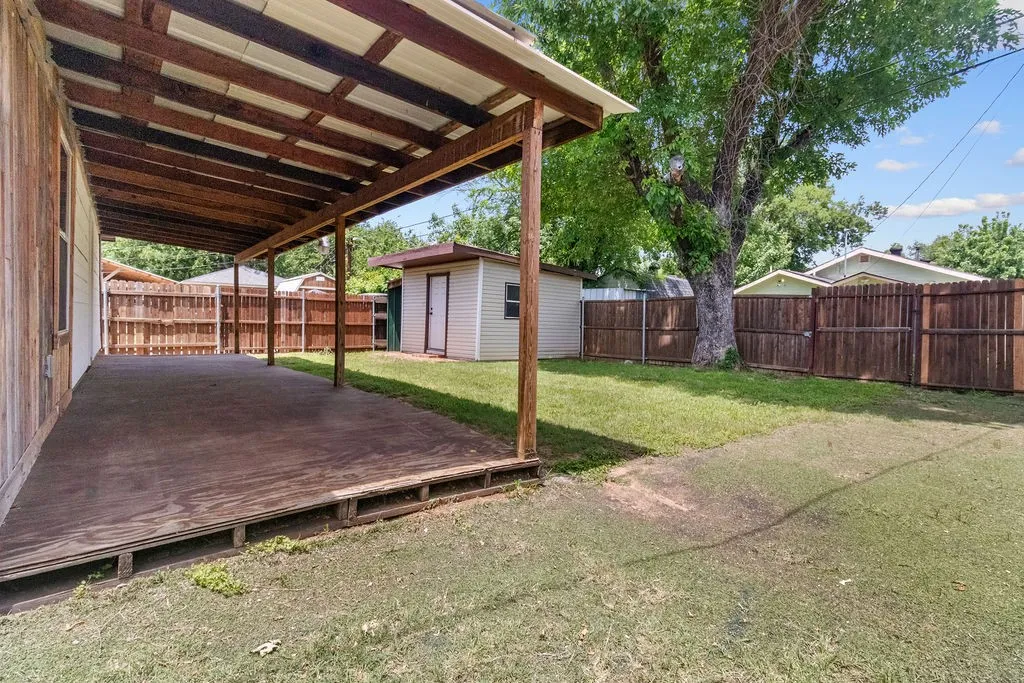 Expansive backyard retreat featuring a wood-finish covered patio with overhead lattice structure, mature shade trees, manicured lawn, wooden fencing, and detached utility structures
