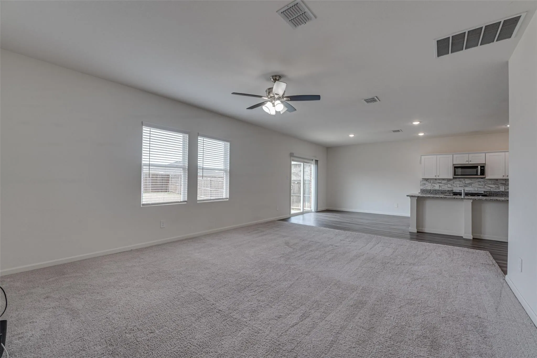Open-concept living space featuring a ceiling fan, dual windows with blinds, a sliding glass door, and wood-finish flooring extending into the kitchen area with white cabinetry and a breakfast bar