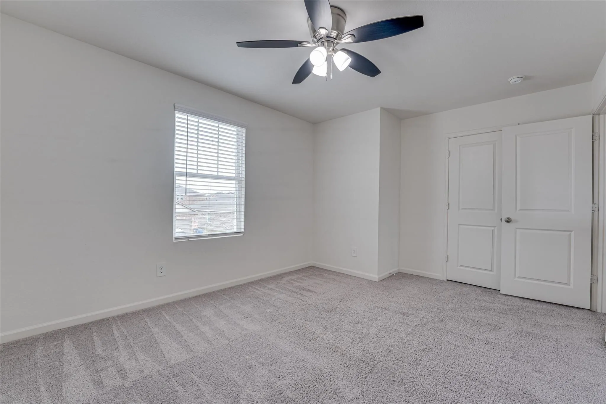 Bright and spacious bedroom featuring neutral wall finishes, carpet flooring with geometric pattern detailing, a ceiling fan with integrated lighting, large window with plantation shutters providing natural light, and dual closets with six-panel​​‌​​​​‌​​‌‌​‌‌​​​‌‌​‌​‌​‌​​​‌​​ doors